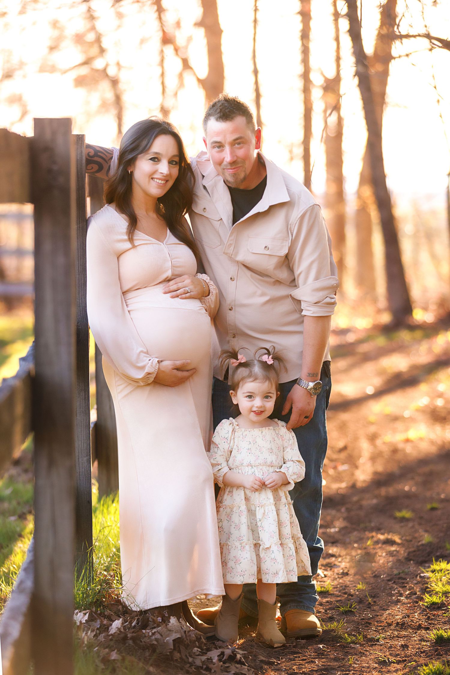 Pregnant woman in blush gown poses with husband and young daughter outdoors during golden hour near a wooden fence.