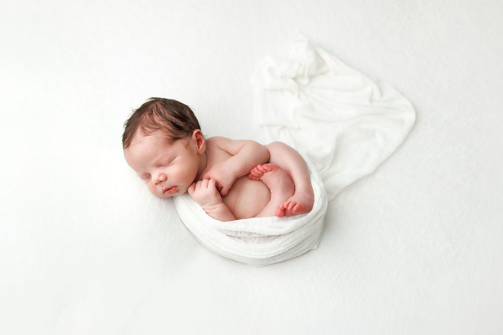 Peaceful newborn baby sleeping curled up in a white bowl wrap on a soft white background.