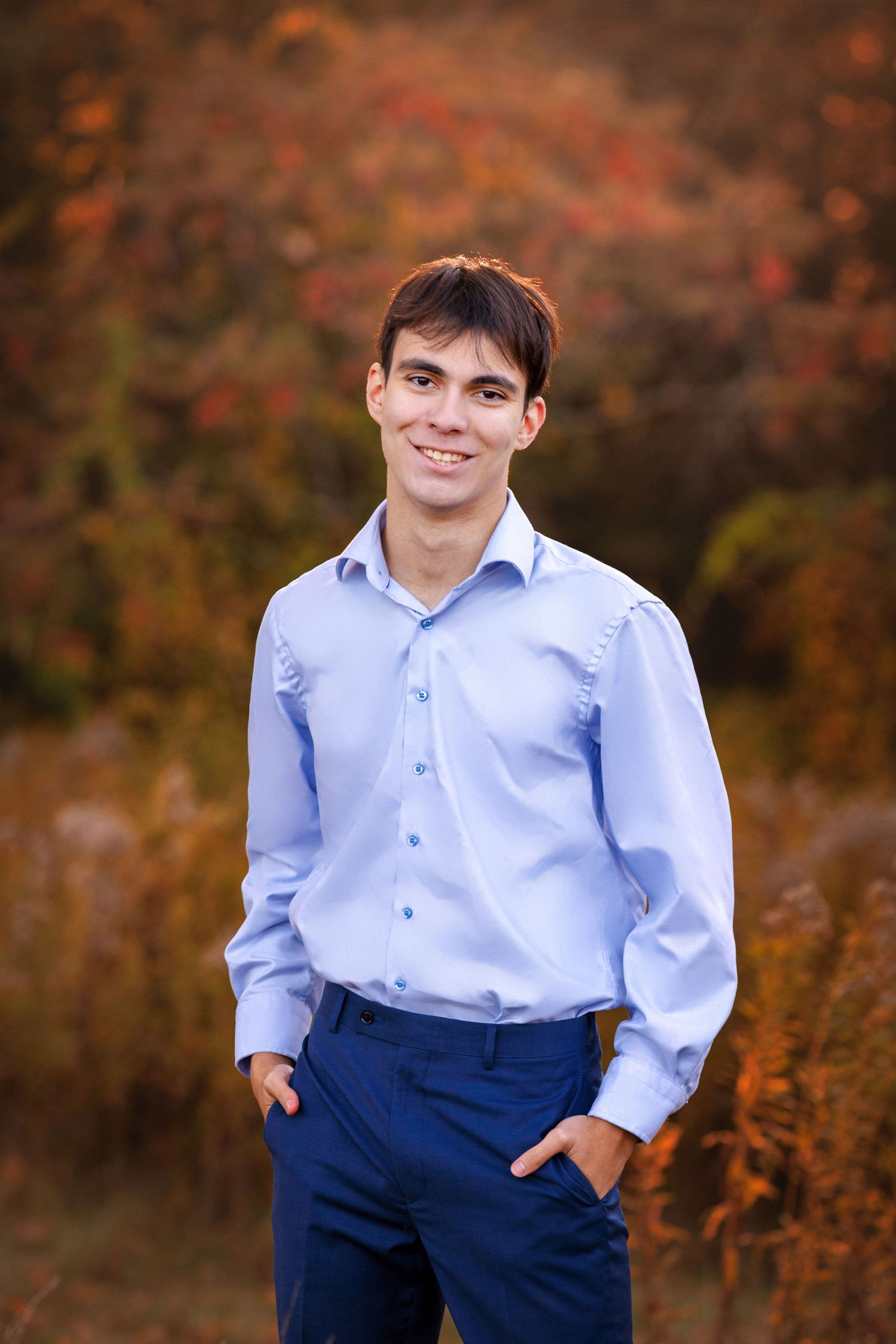 Young man in blue shirt and navy pants smiling outdoors with colorful autumn foliage background.