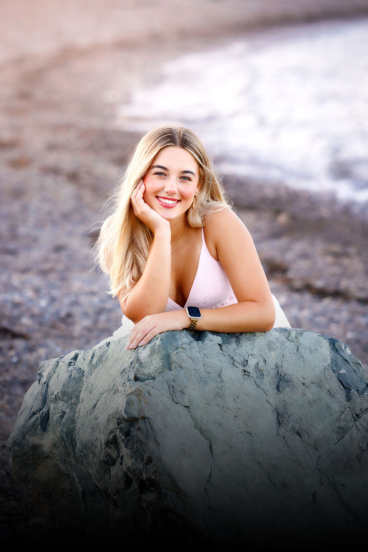 Smiling blonde woman leaning on a rock at the beach, wearing a white top and smartwatch.