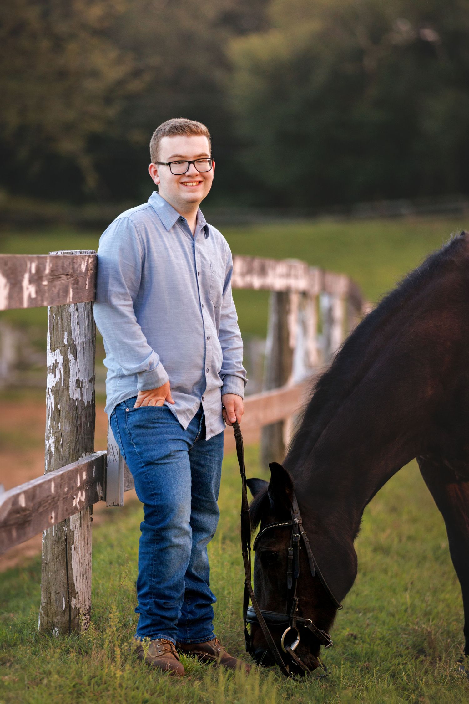 Young man with glasses smiles leaning on a wooden fence while holding a dark horse grazing on a green farm.