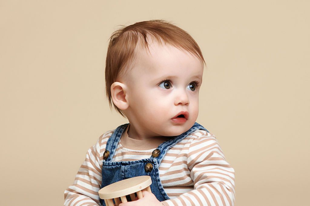 Adorable baby in denim overalls and striped shirt holding a wooden toy against a beige background.