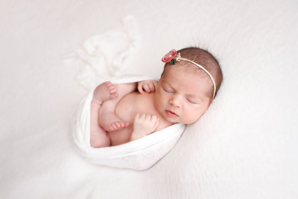Peaceful newborn baby girl sleeping curled up in white wrap with a delicate floral headband.