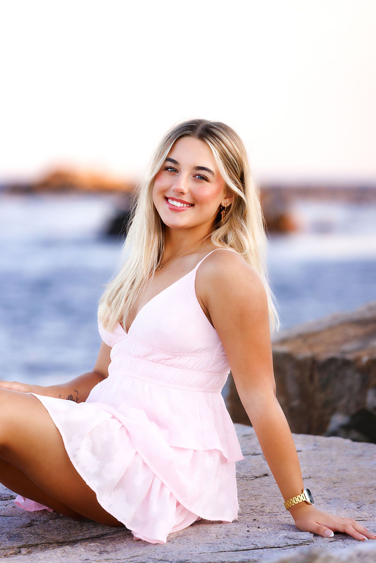 Windham NH Senior in a light pink sundress poses gracefully on coastal rocks by the ocean at sunset.