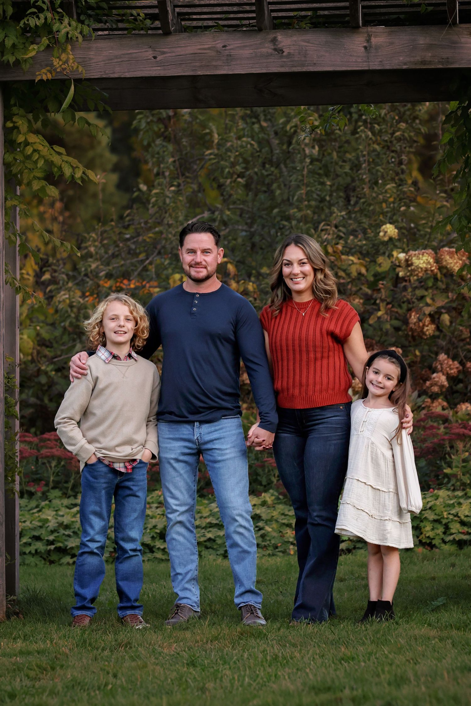 A family of four stands together on grass in autumn, wearing casual clothing against a backdrop of fall foliage.