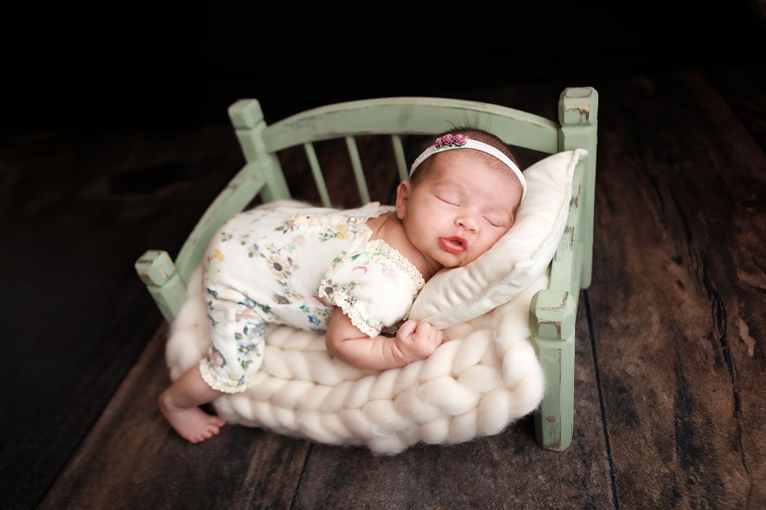 Newborn baby sleeping peacefully in a miniature green wooden bed with a floral onesie and pink headband.