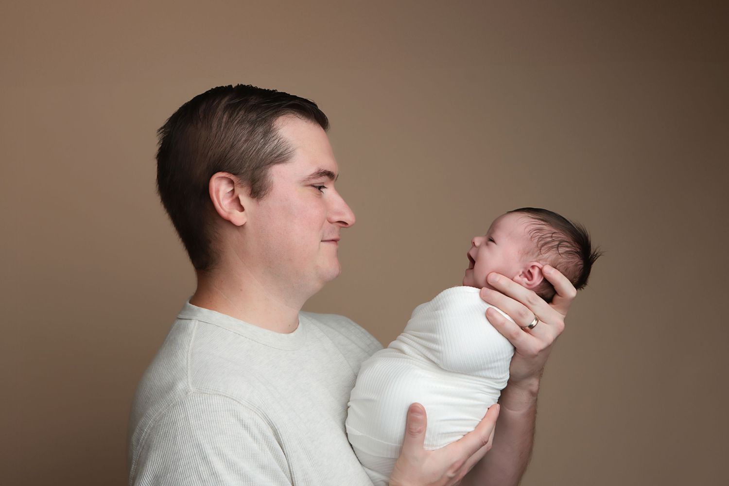 A parent in a white sweater holds and lovingly gazes at their newborn baby in a tender moment against a brown background.