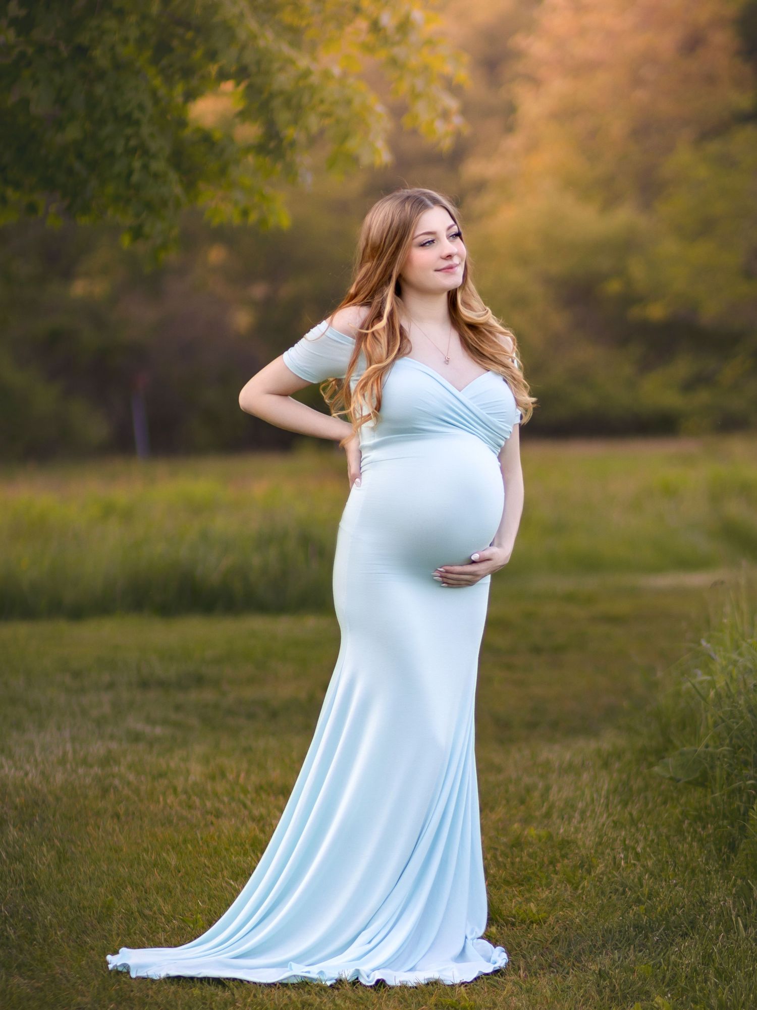 A woman in a flowing light blue dress poses gracefully in a sunlit autumn field during a maternity photoshoot.