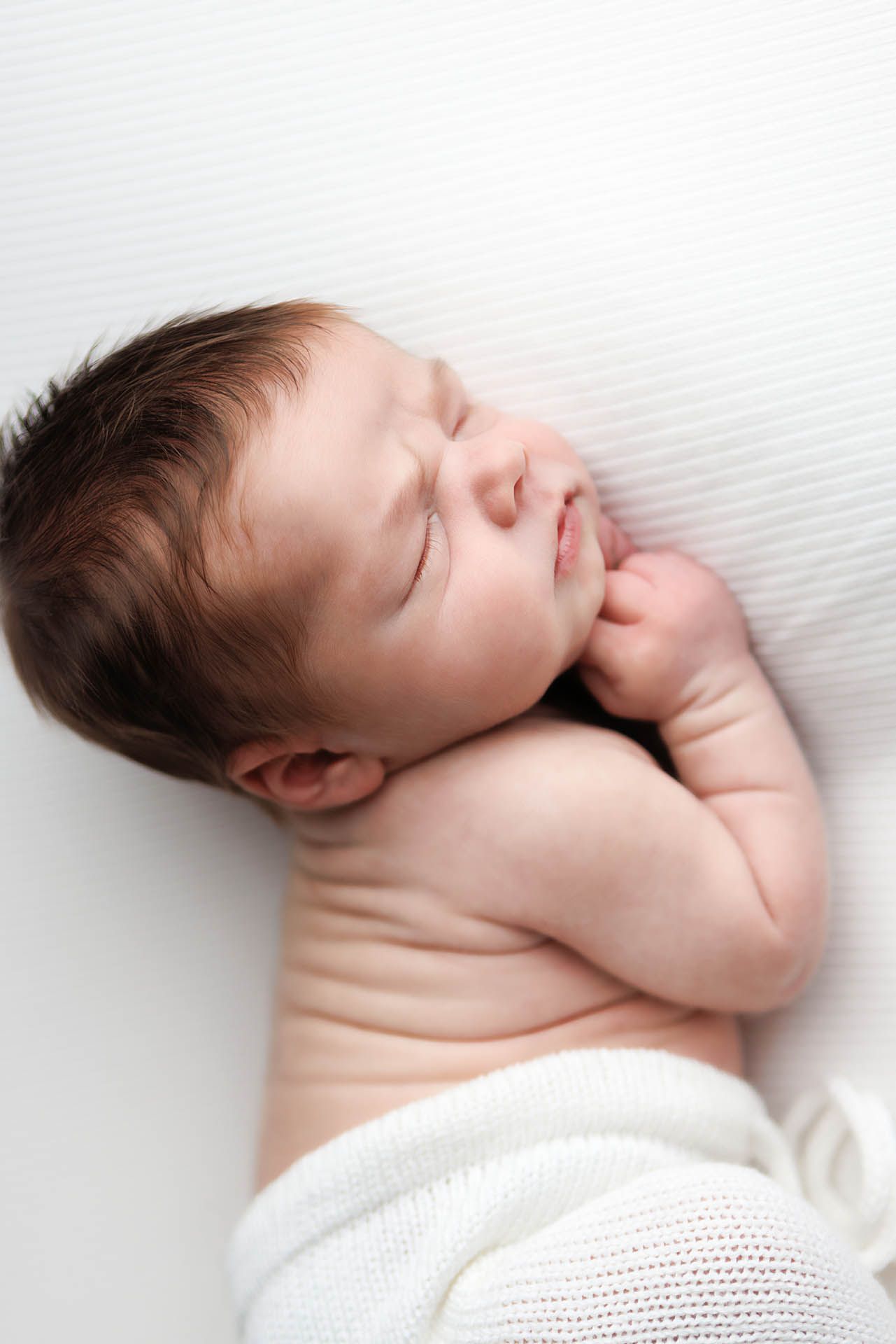 Peaceful newborn baby sleeping on white blanket with dark hair and tiny hands tucked under chin.