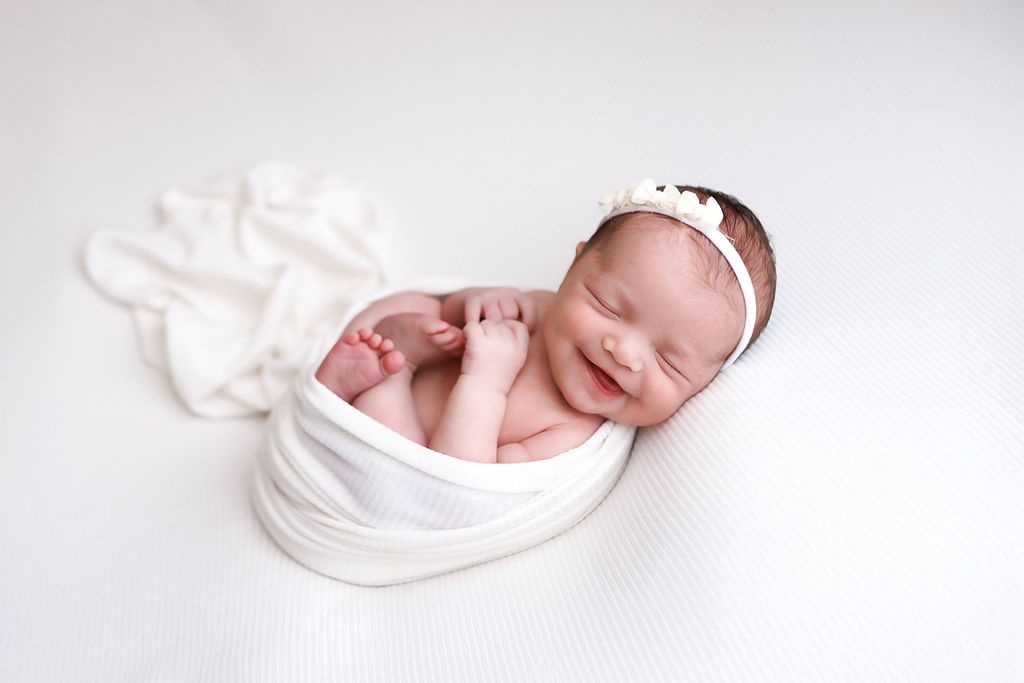 Smiling newborn baby wrapped in white swaddle cloth wearing a delicate white headband on a white background.