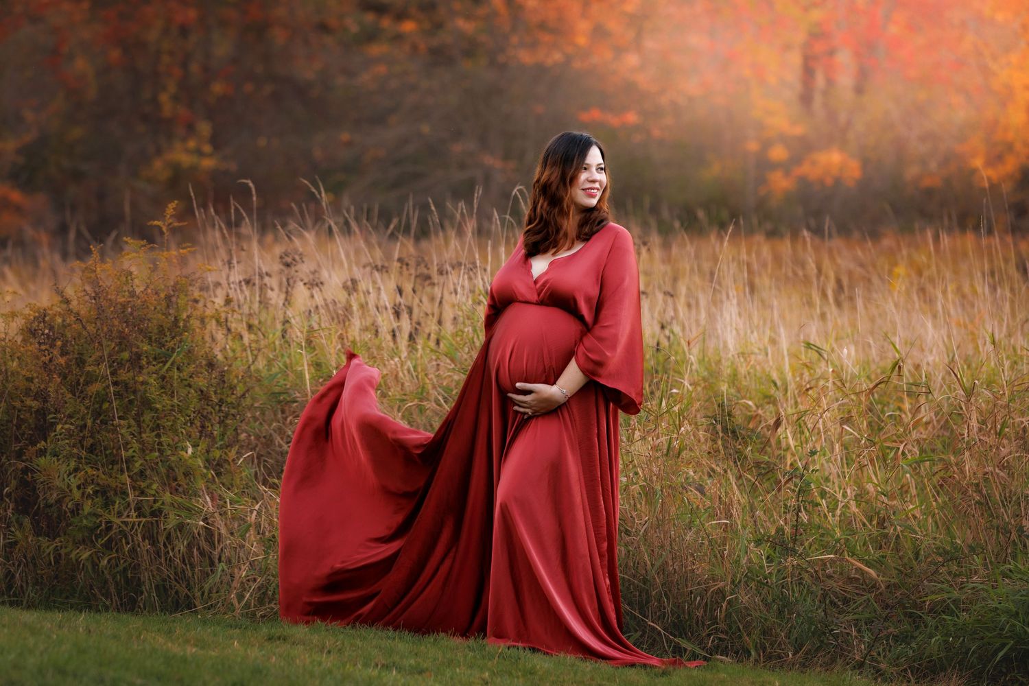A figure in a flowing red dress poses gracefully in an autumn field during golden hour sunset. NH Maternity Portrait.