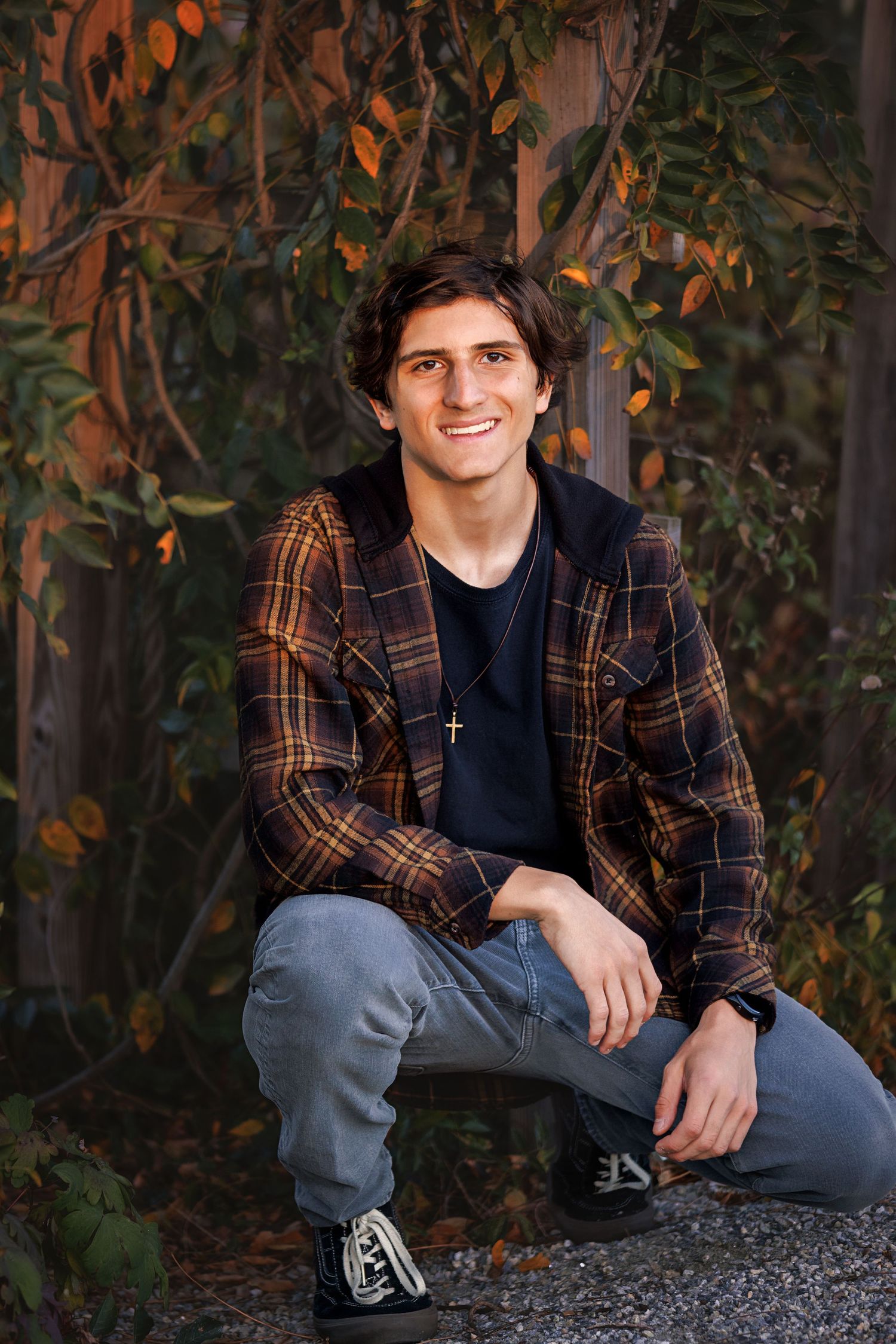 Senior in brown plaid jacket and jeans poses outdoors among autumn foliage and trees.