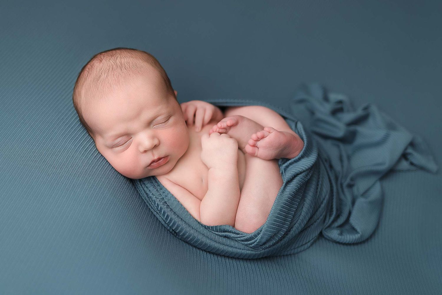 Newborn baby sleeping peacefully on soft teal fabric in artistic curled pose during professional photo session.