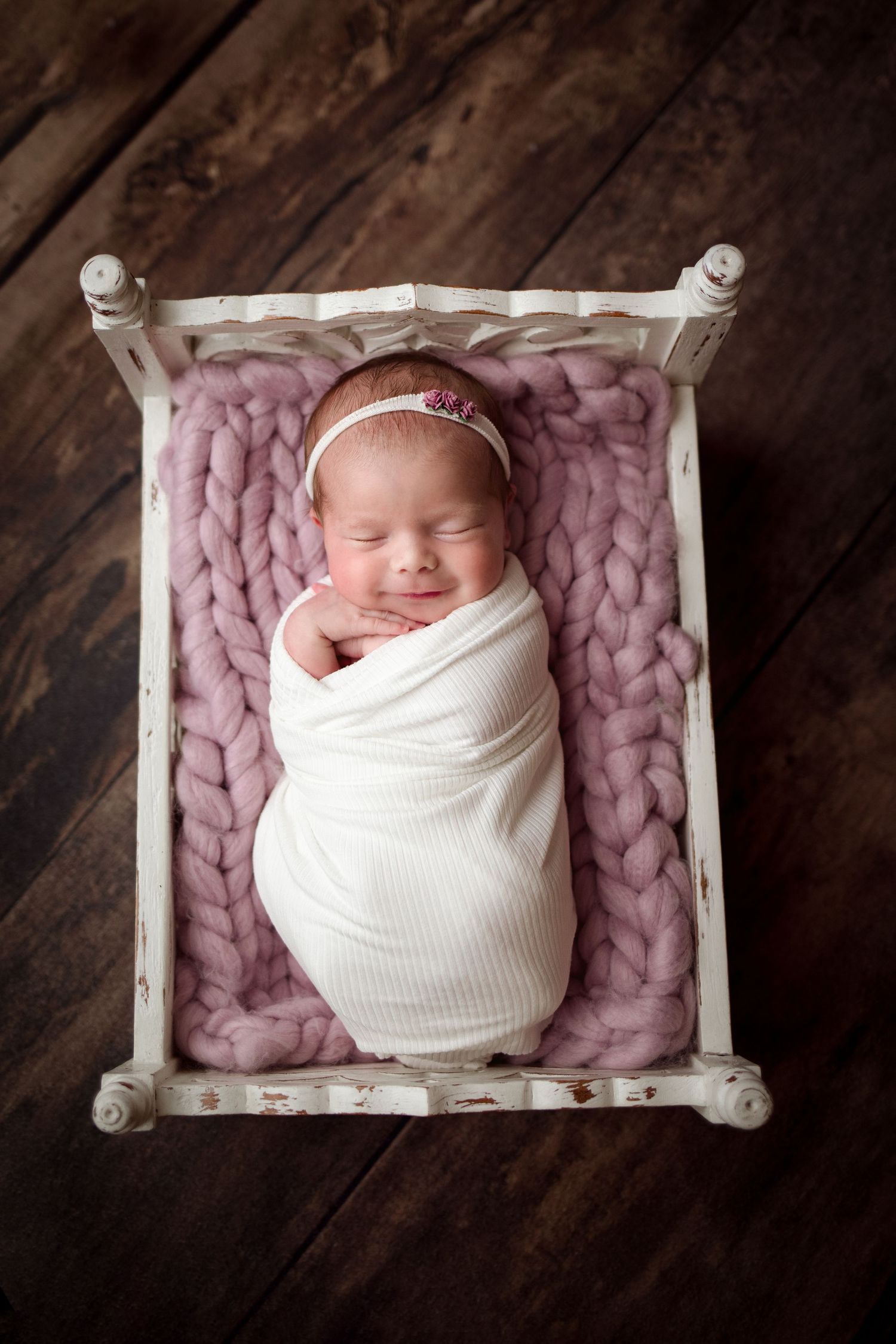 A sleeping newborn swaddled in white fabric rests peacefully on a chunky knit pink blanket in a rustic wooden setting.