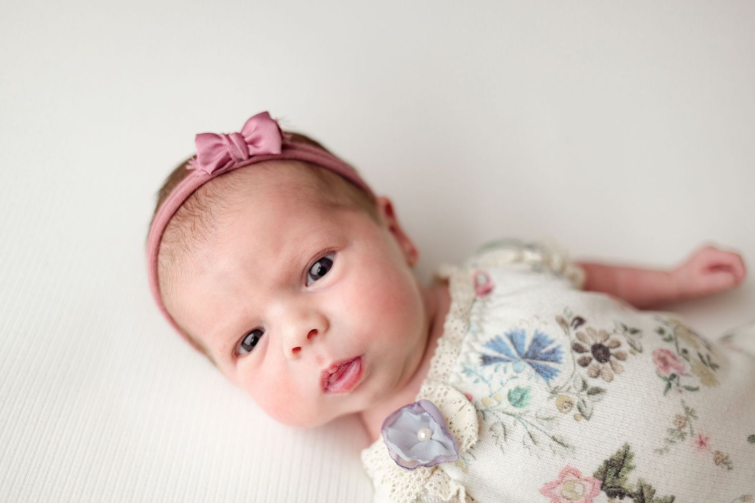 A newborn baby wearing a floral outfit and pink bow headband lies on white bedding during a professional photo session.