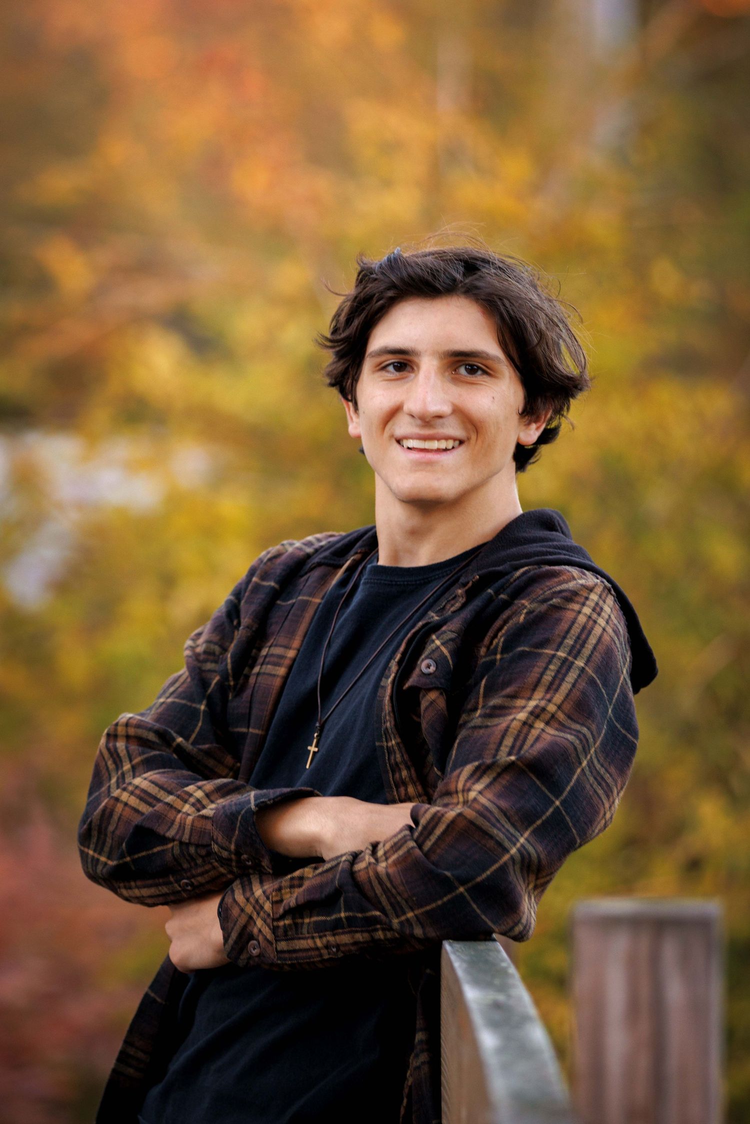 High School Senior in dark plaid jacket poses with crossed arms against blurred fall foliage background.