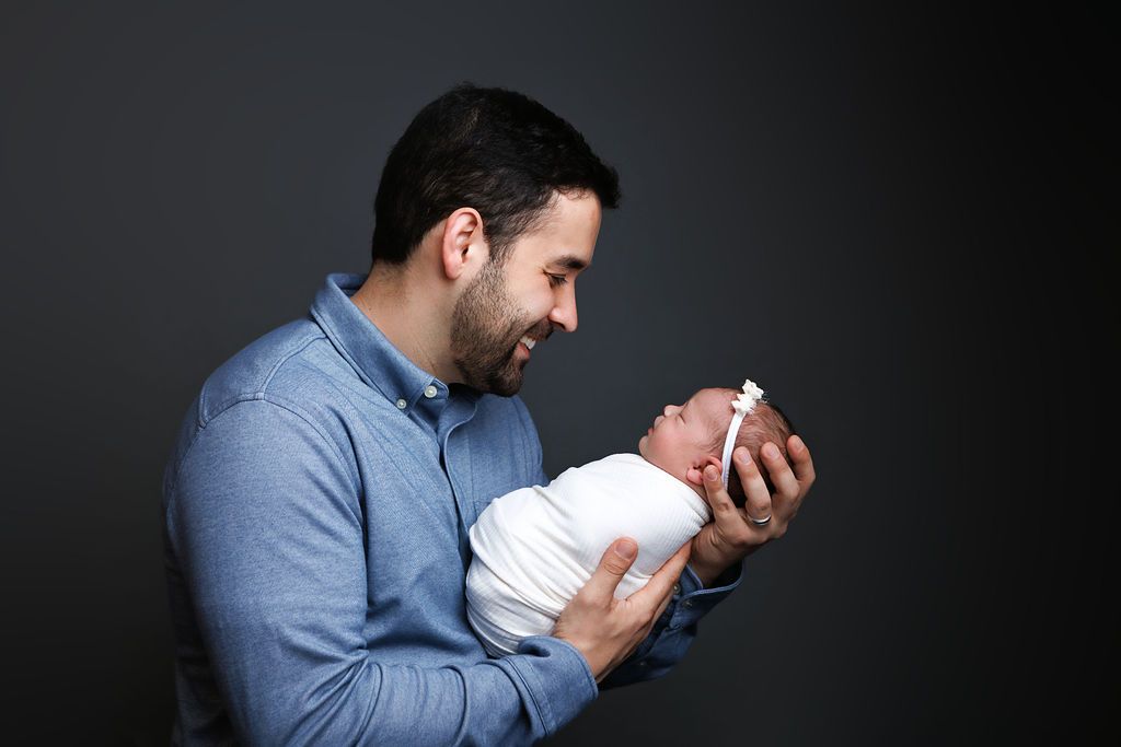 A father tenderly holds his newborn baby in his arms, smiling lovingly against a dark background.