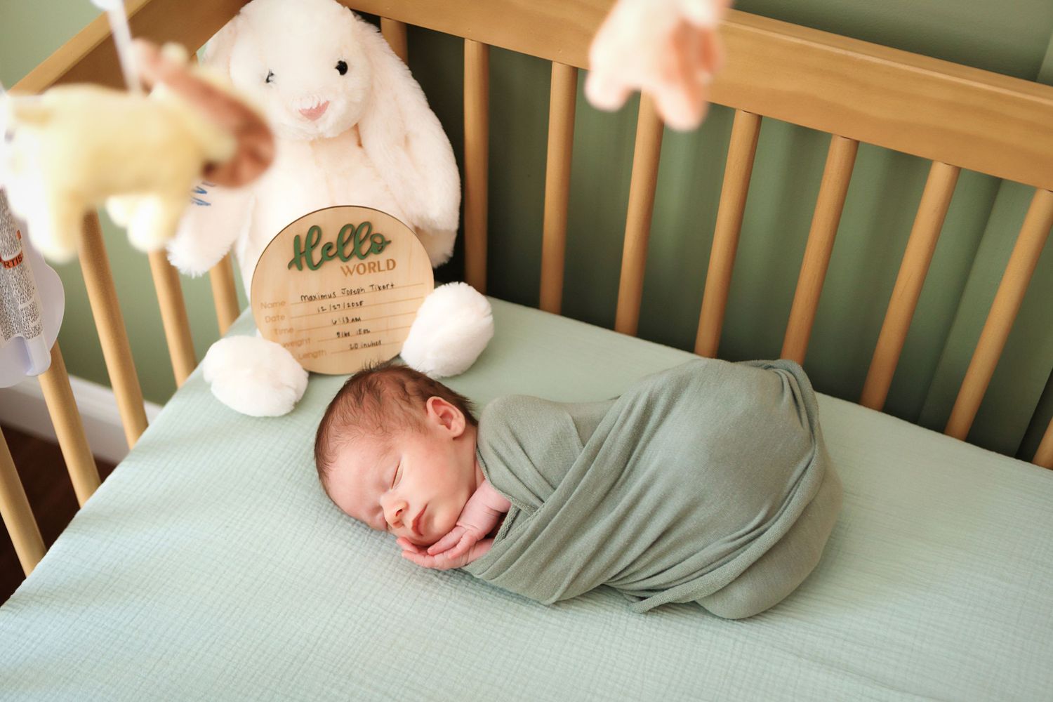 Newborn baby swaddled in sage green wrap sleeping peacefully in wooden crib with bunny stuffed animal and Hello birth sign.