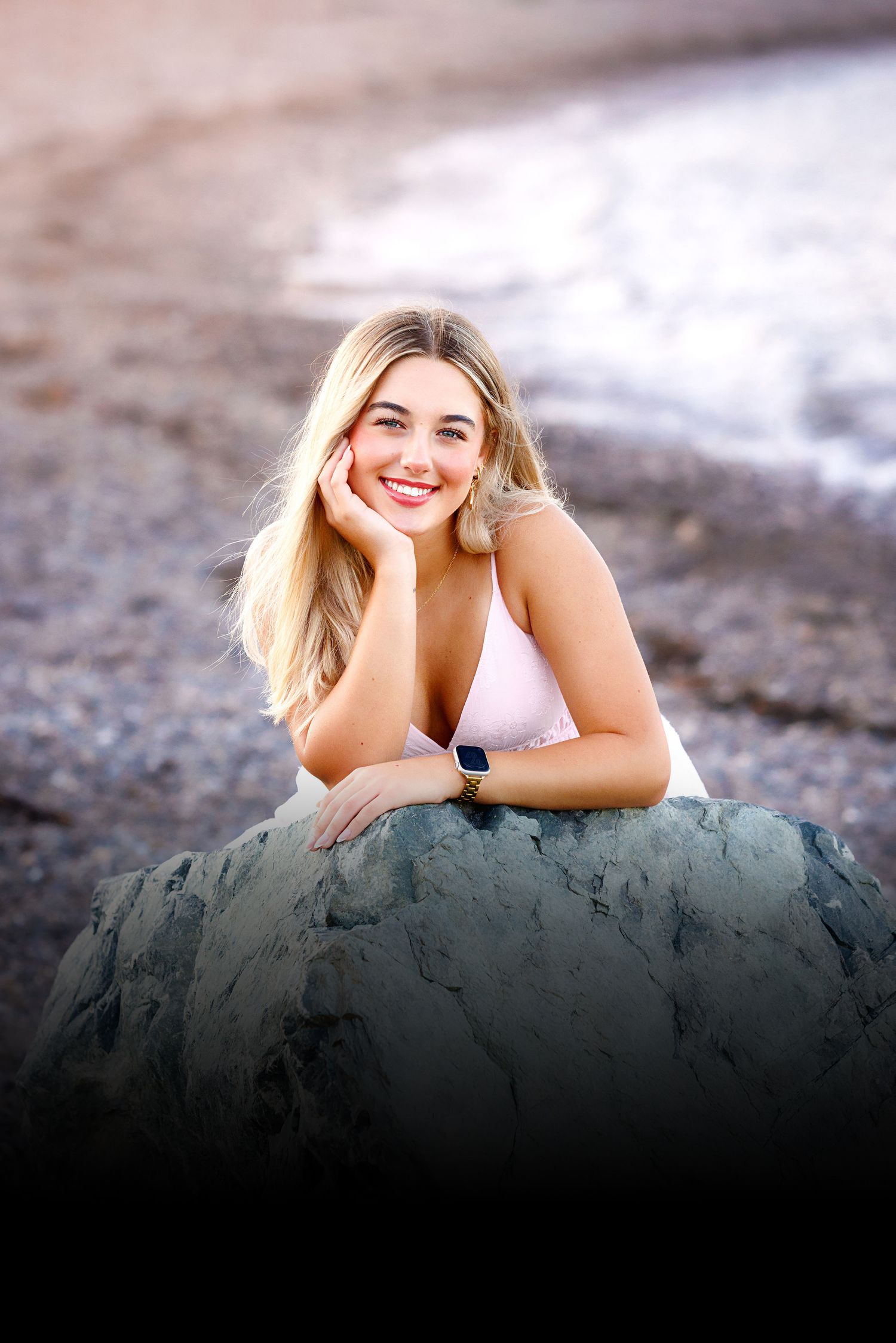 Smiling blonde woman leaning on a rock at the beach, wearing a white top and smartwatch.