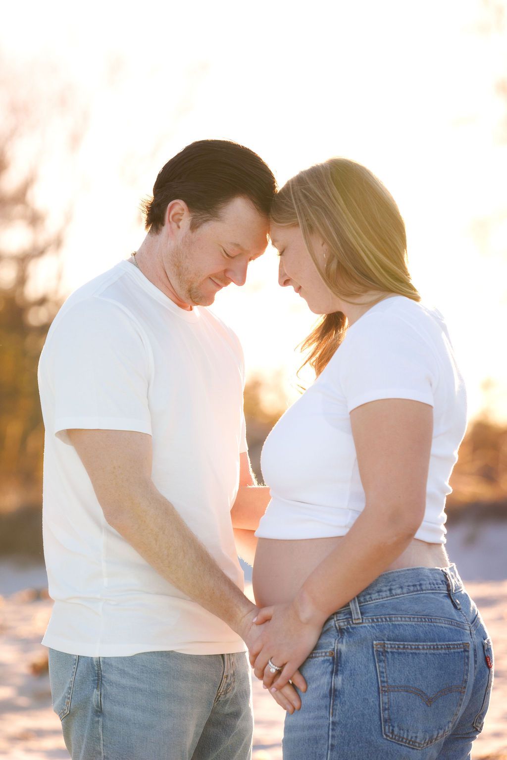Couple in white shirts touching foreheads, hands on pregnant belly, glowing sunset backdrop outdoors.