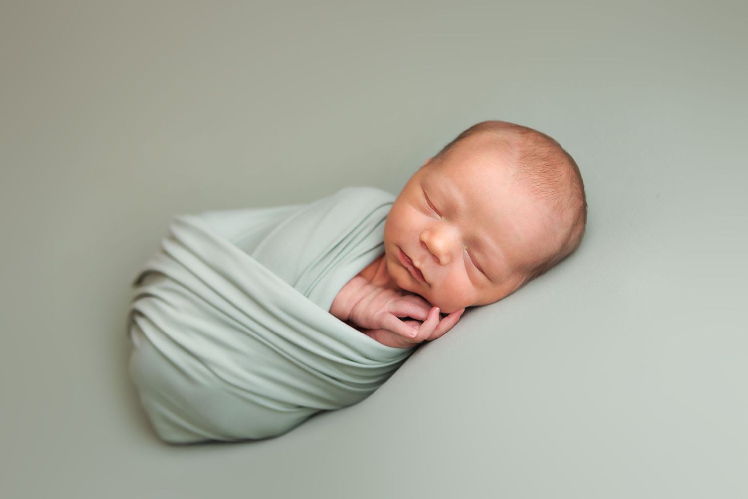 Peaceful newborn baby swaddled in a soft mint green wrap, sleeping serenely against a matching green backdrop.