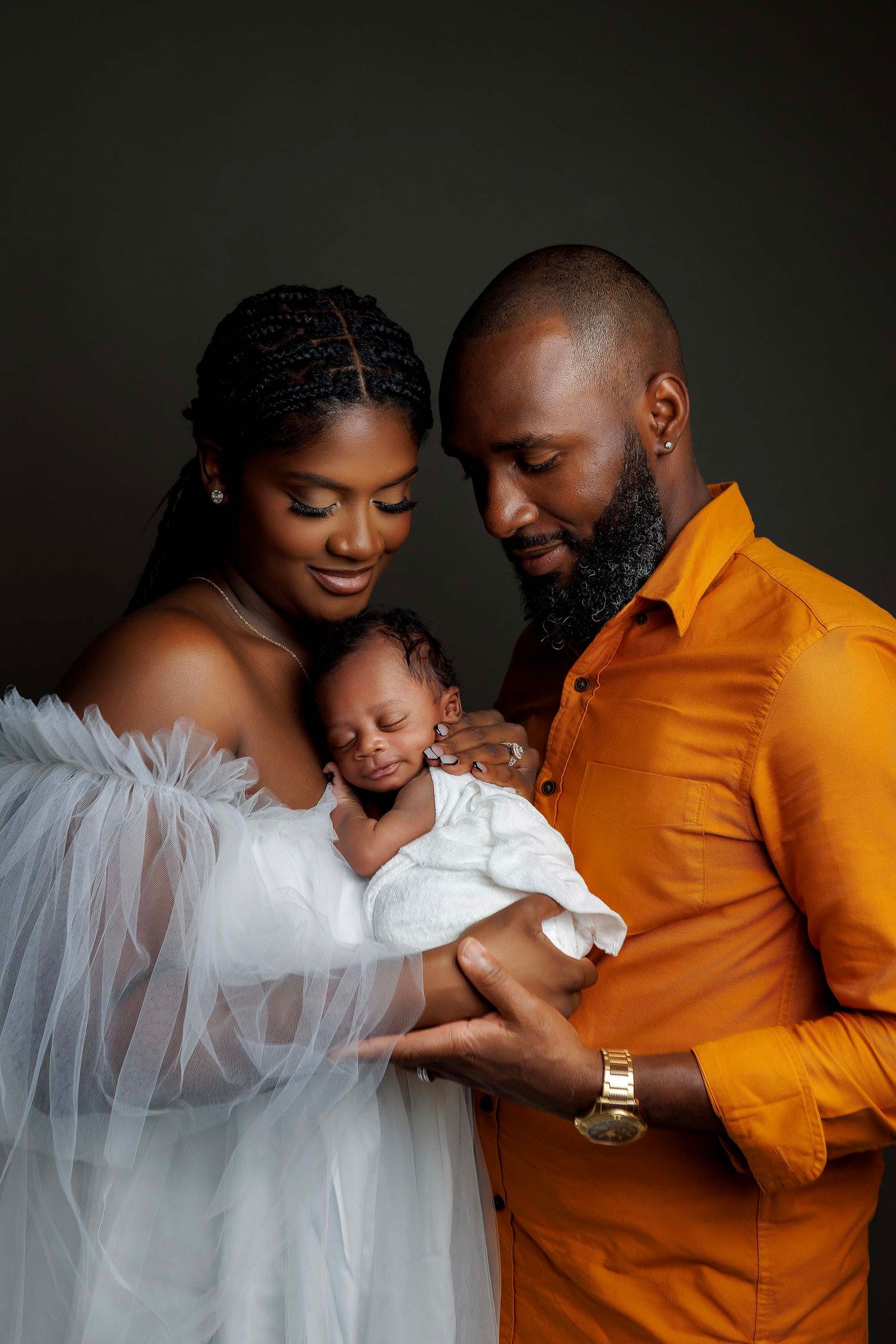 A tender family portrait against dark backdrop shows parents cradling their sleeping newborn baby in white clothing.