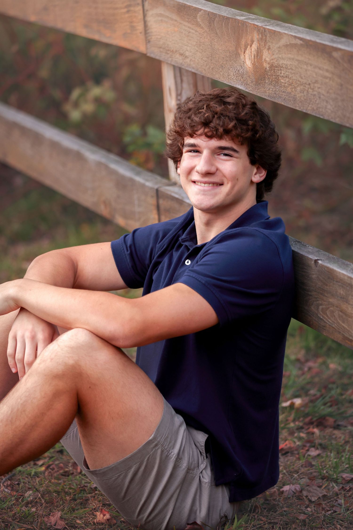 High School Senior in navy shirt and gray shorts sitting casually against wooden fence outdoors.