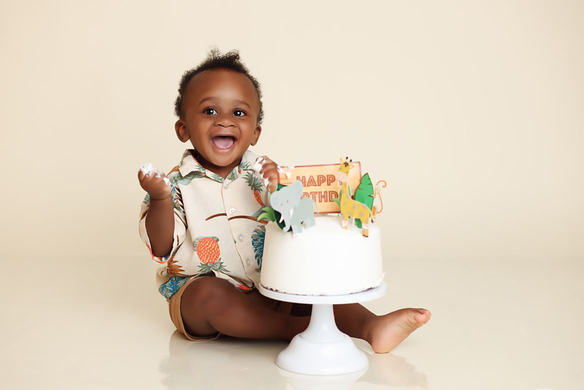 A smiling baby in a white outfit sits next to a white cake stand with a birthday cake.