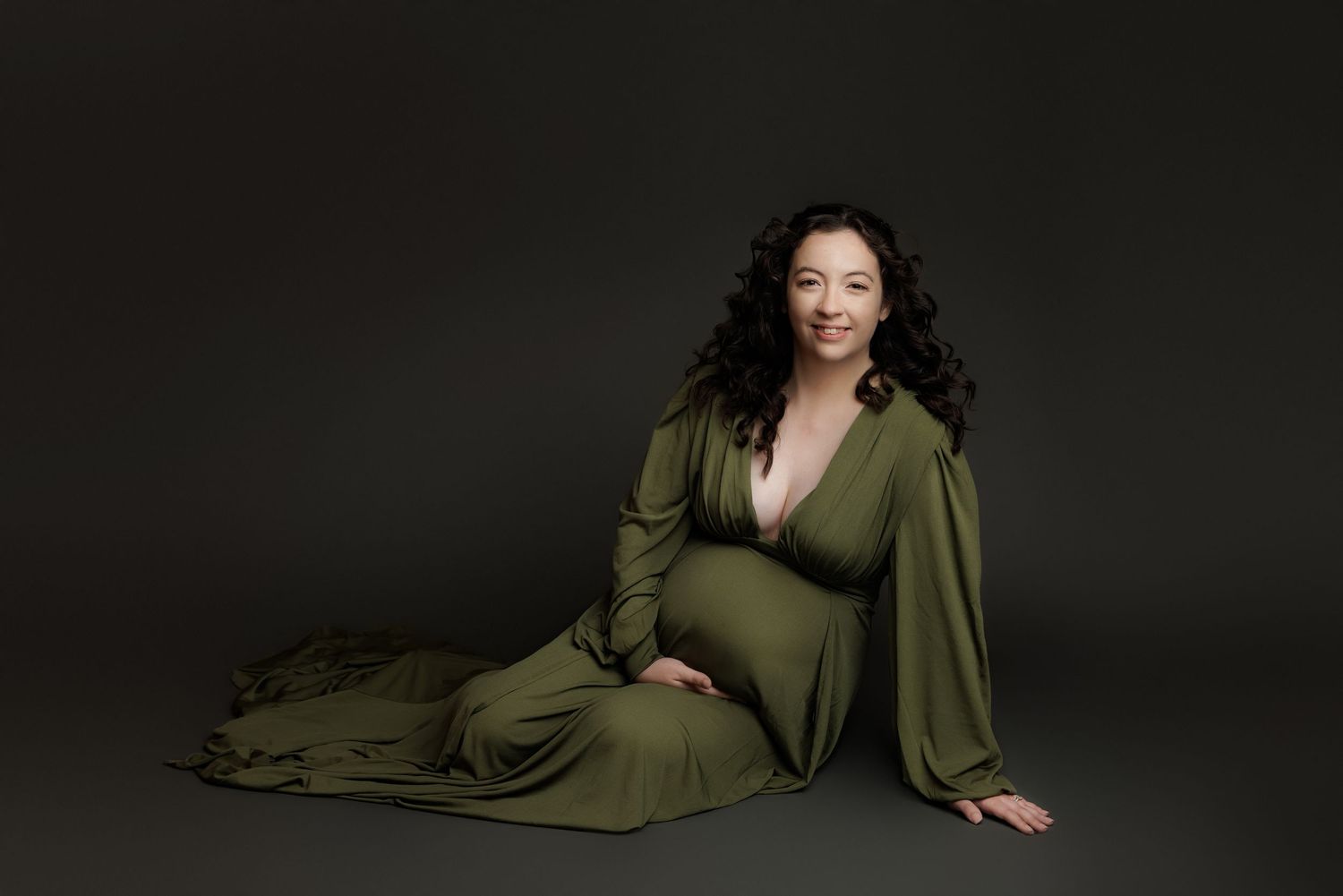 Elegant maternity portrait featuring a flowing olive green dress against a dark studio backdrop.