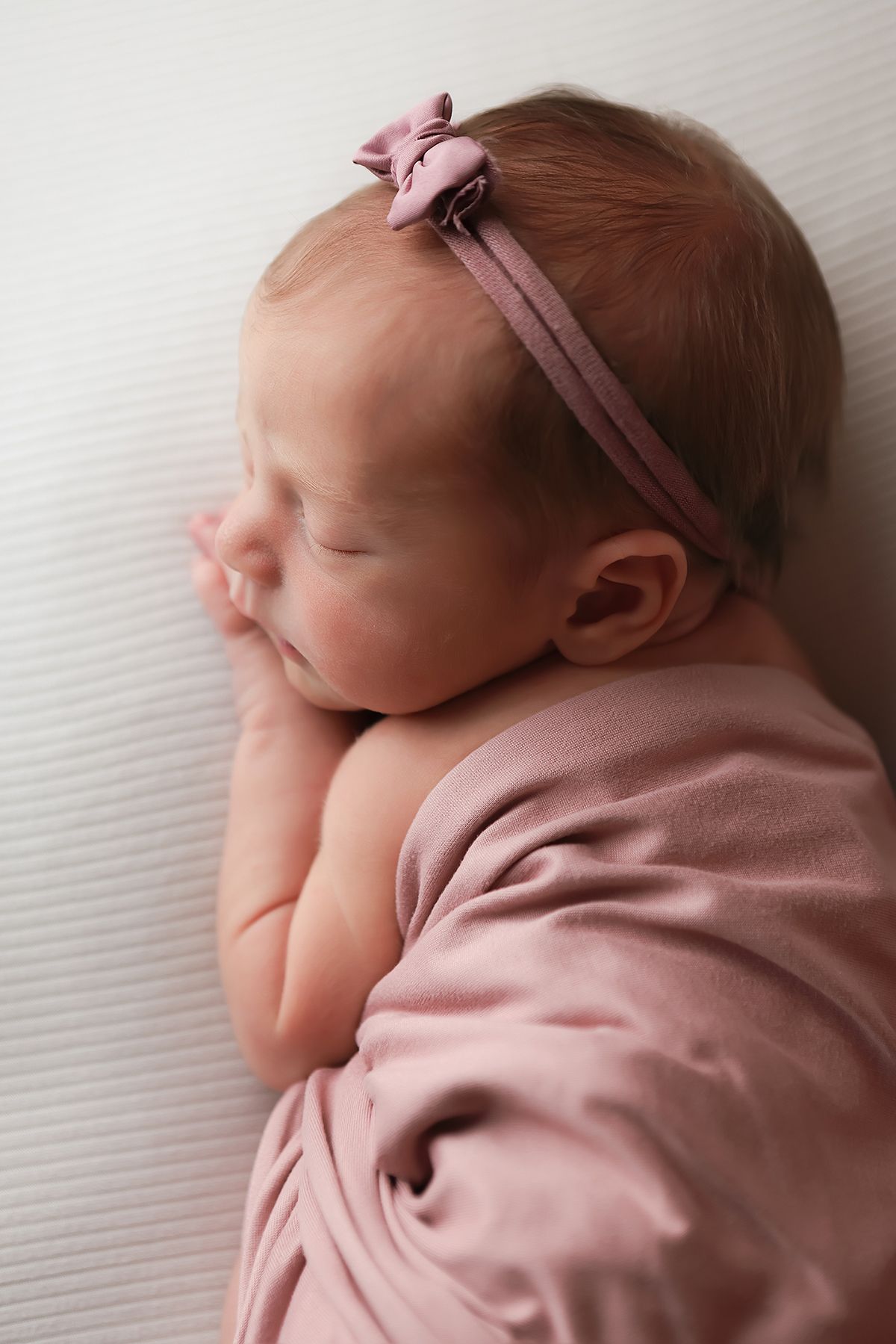 Newborn baby wrapped in soft pink swaddle wearing matching headband rests peacefully against white textured background.