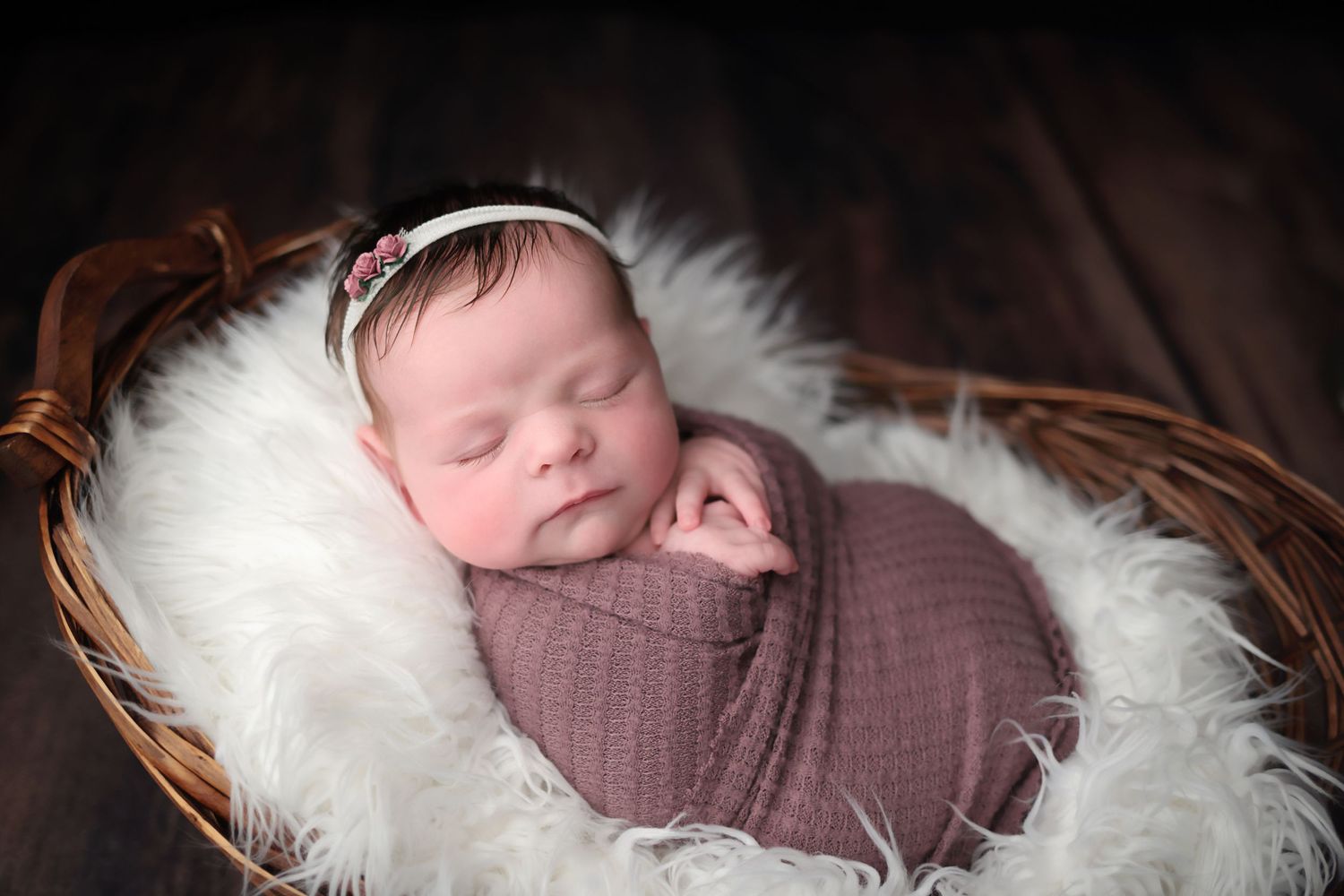 A sleeping baby wrapped in a pink swaddle blanket lies peacefully in a white fur-lined basket.