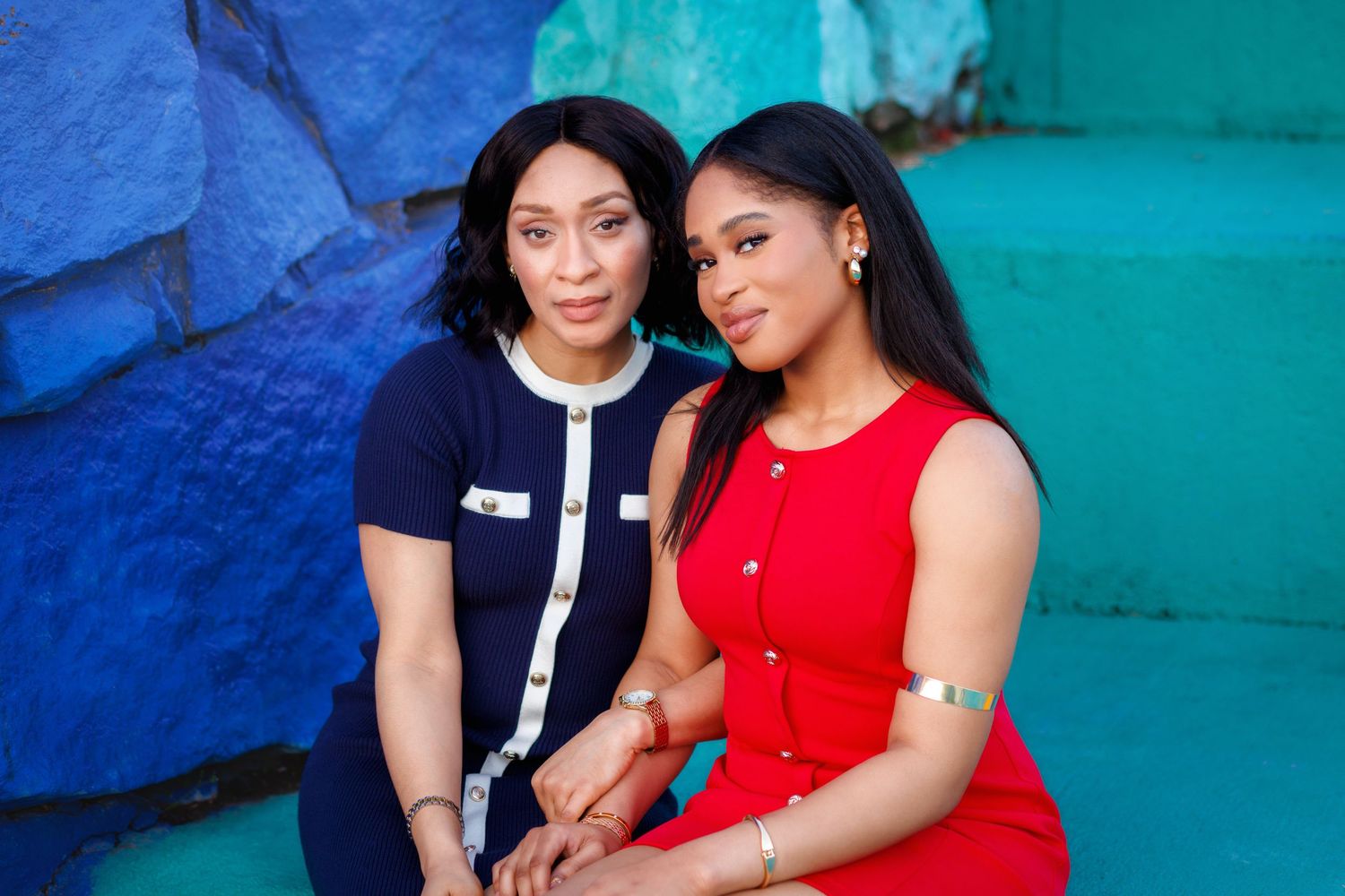 Two women pose together against a vibrant blue wall, one in a navy dress and one in a red dress.