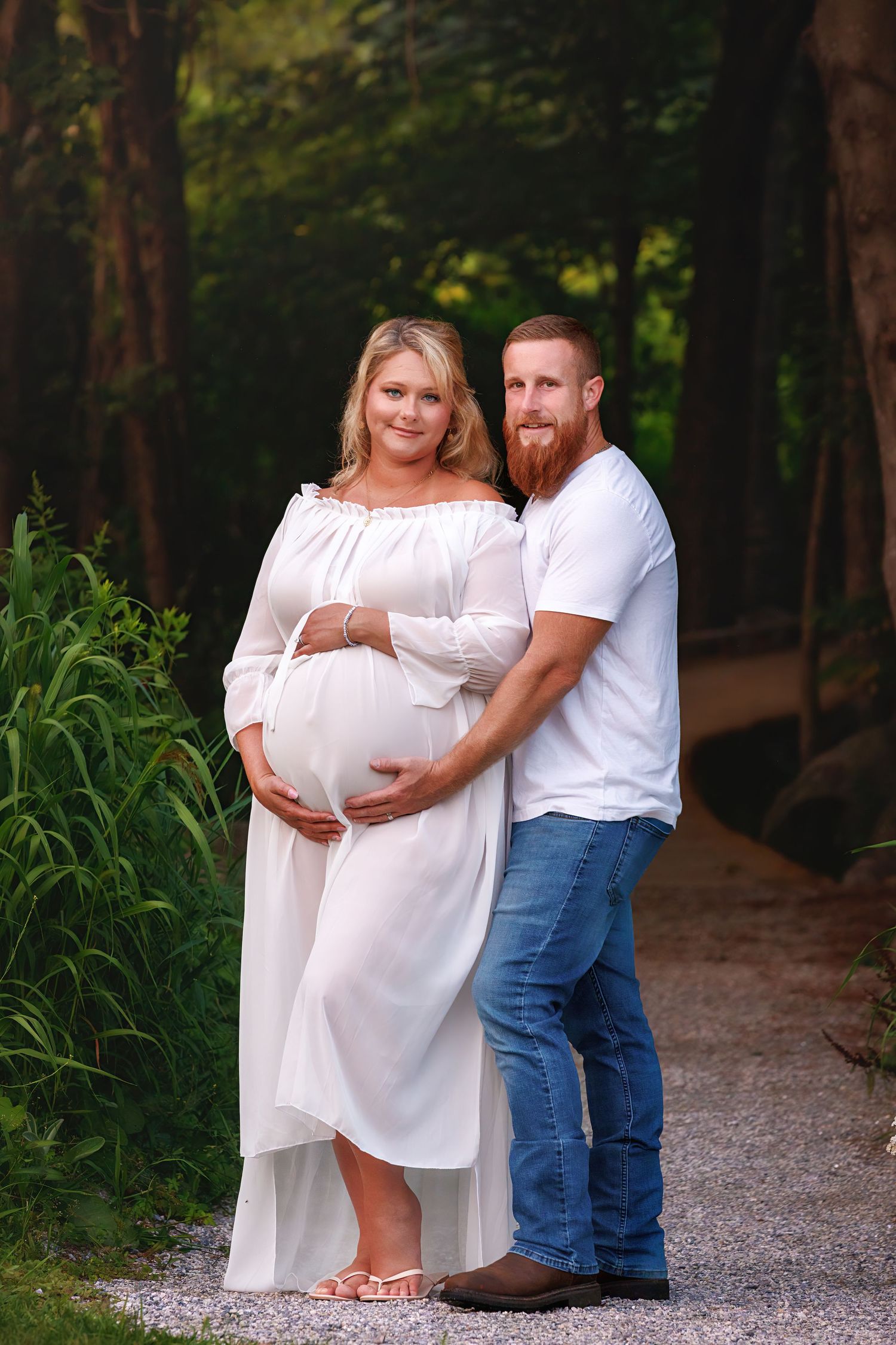 Couple in white outfits posing for maternity photos on a gravel path surrounded by lush greenery.
