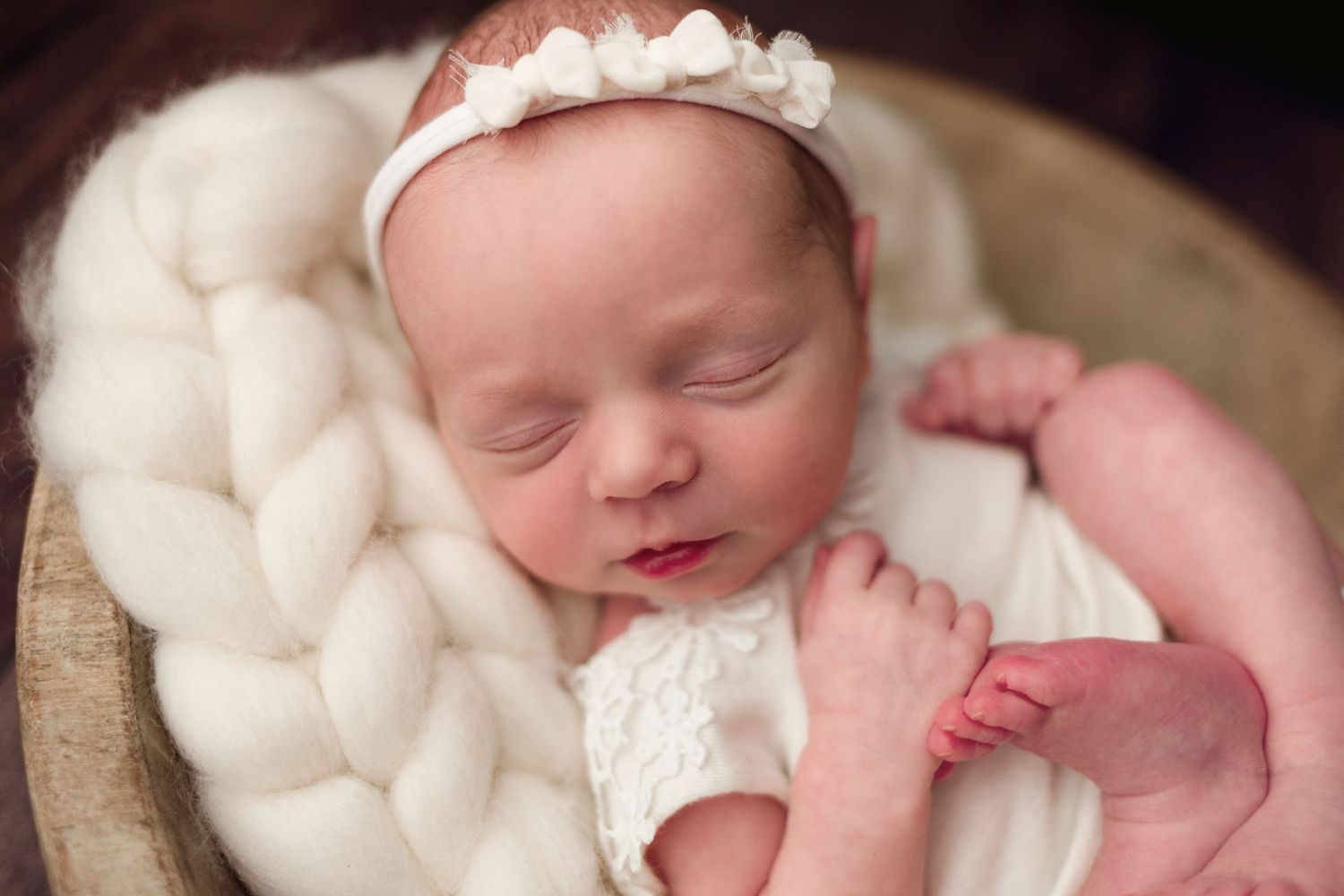 Newborn baby sleeping peacefully on white knitted blanket wearing delicate lace headband and dress.