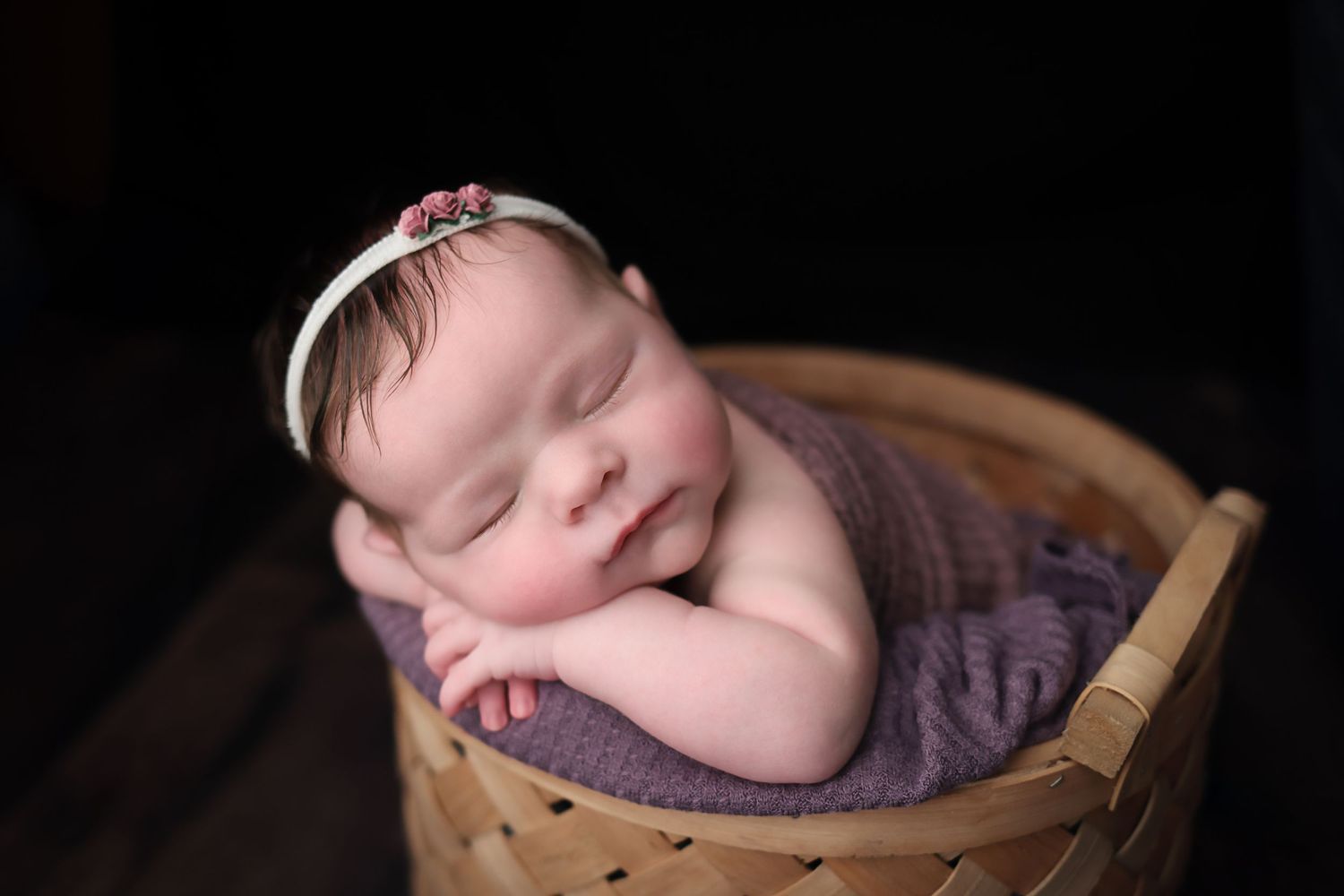 A sleeping newborn baby rests peacefully in a wooden bowl with purple fabric padding.