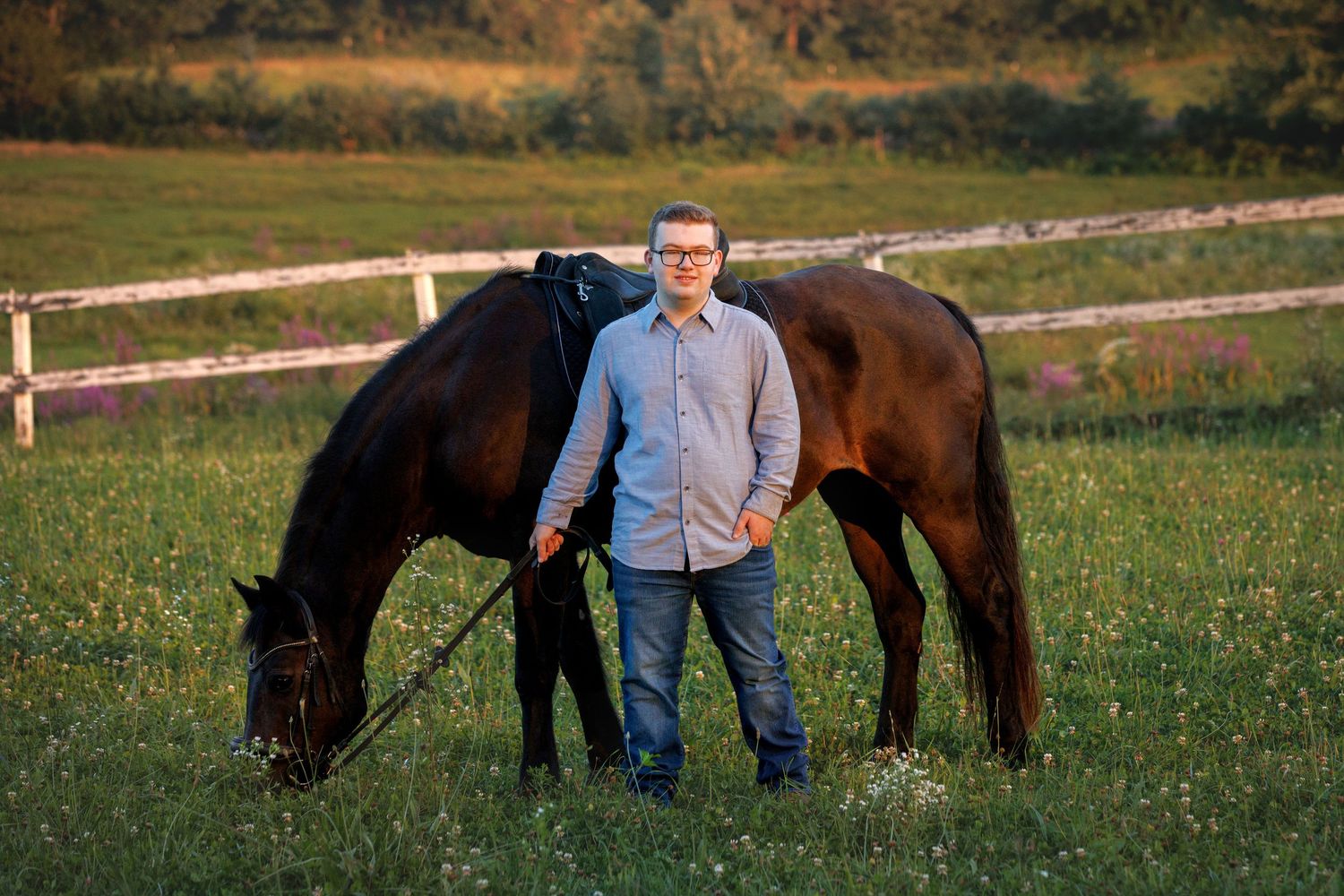Young man in blue shirt and jeans standing with a dark bay horse grazing in a green pasture.