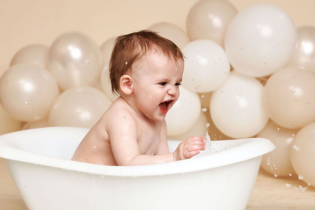 Happy laughing baby splashing in a white mini bathtub with water droplets and neutral balloons behind.