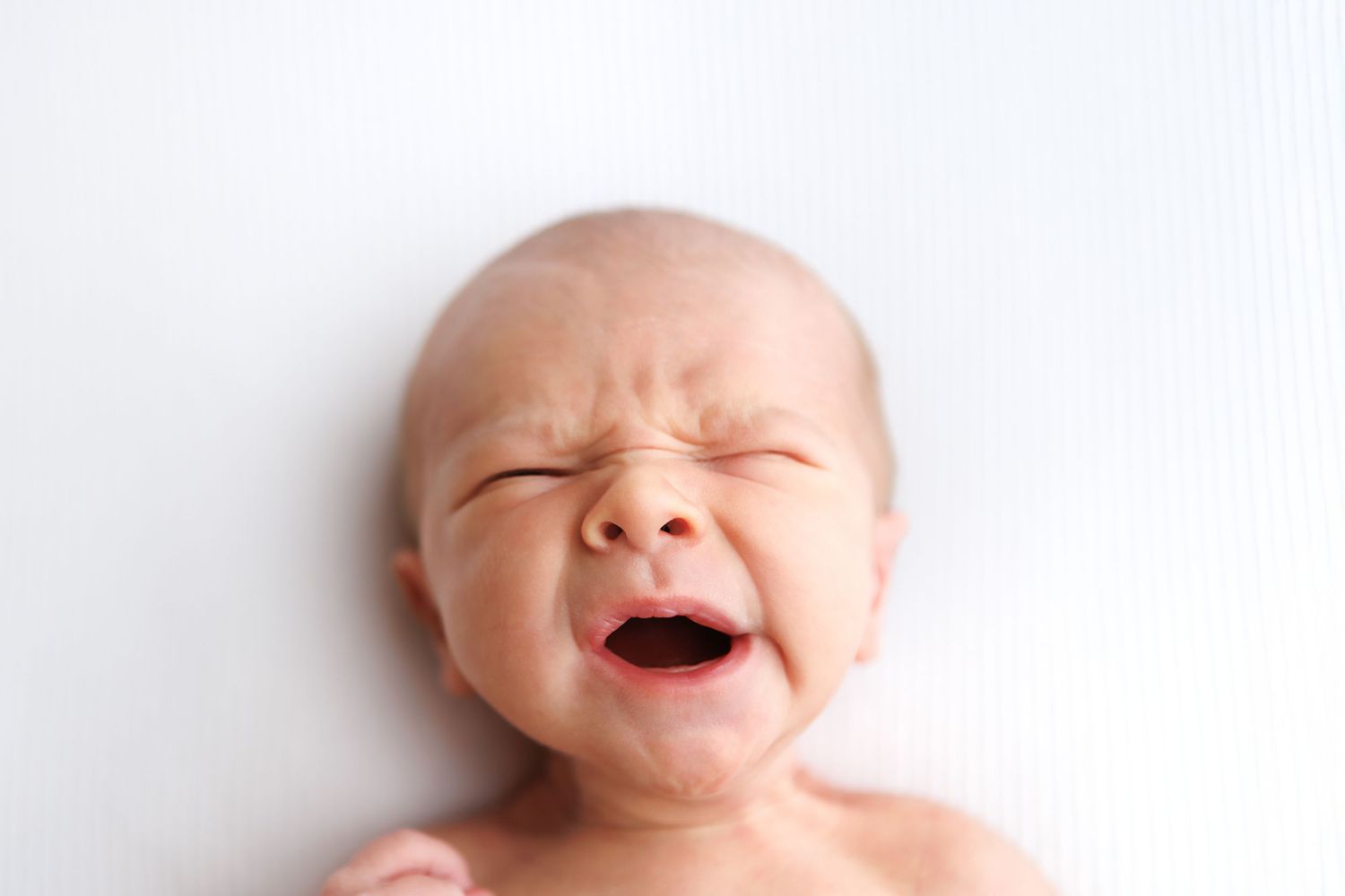 Newborn baby crying with eyes closed, lying on white background, close-up portrait of infant distress.