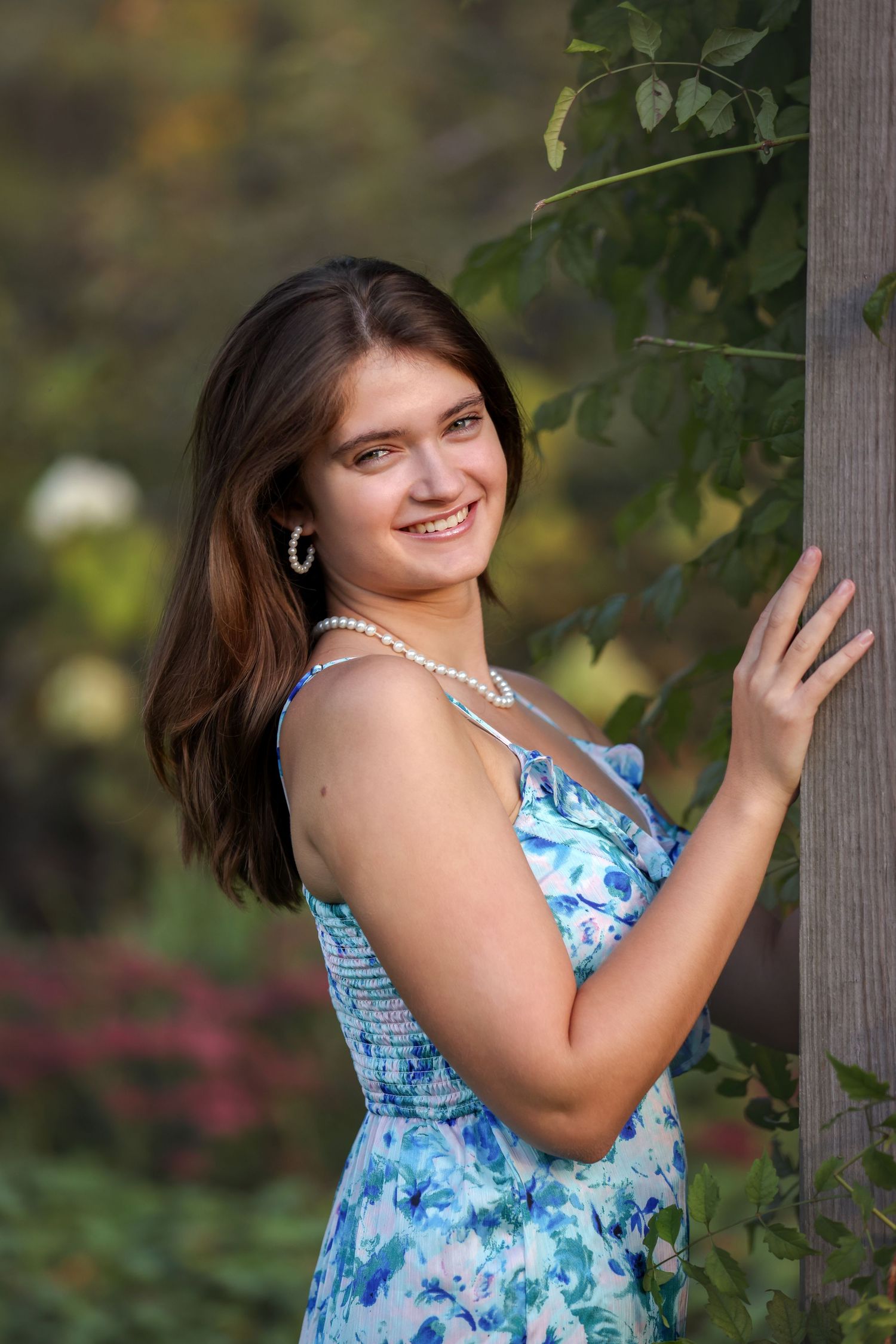 Young woman in floral blue dress and pearl necklace smiles outdoors near wooden post with green foliage background.