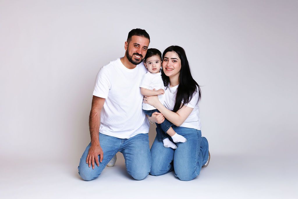 Happy family portrait with parents in white t-shirts and jeans sitting on floor holding their baby on white background.