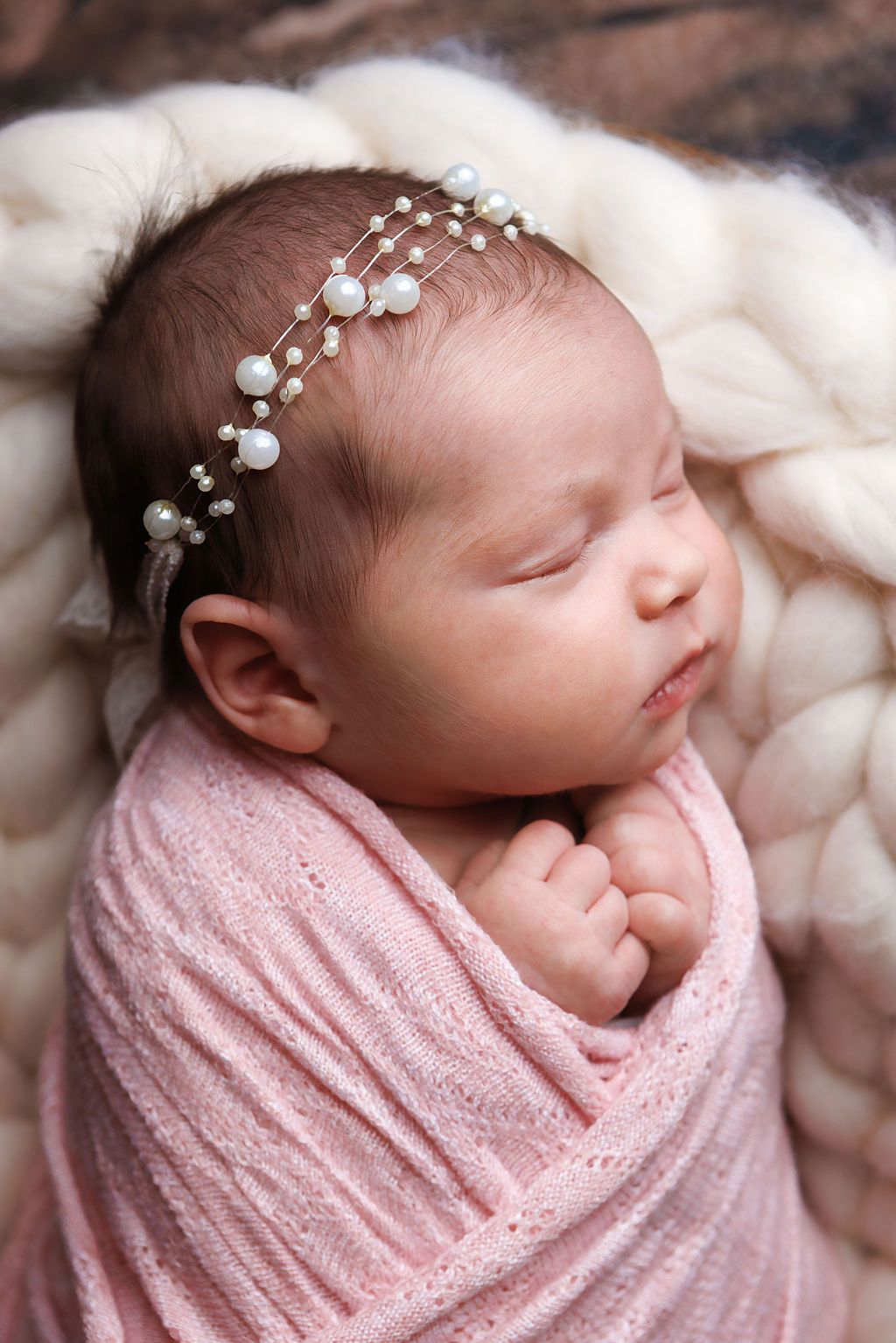 Sleeping newborn baby girl swaddled in pink wrap wearing delicate pearl headband on soft cream blanket.