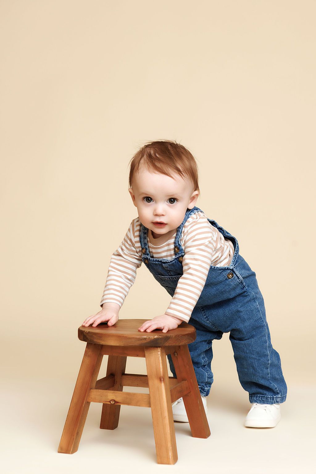 Adorable baby in denim overalls and striped shirt standing at a small wooden stool on a beige background.