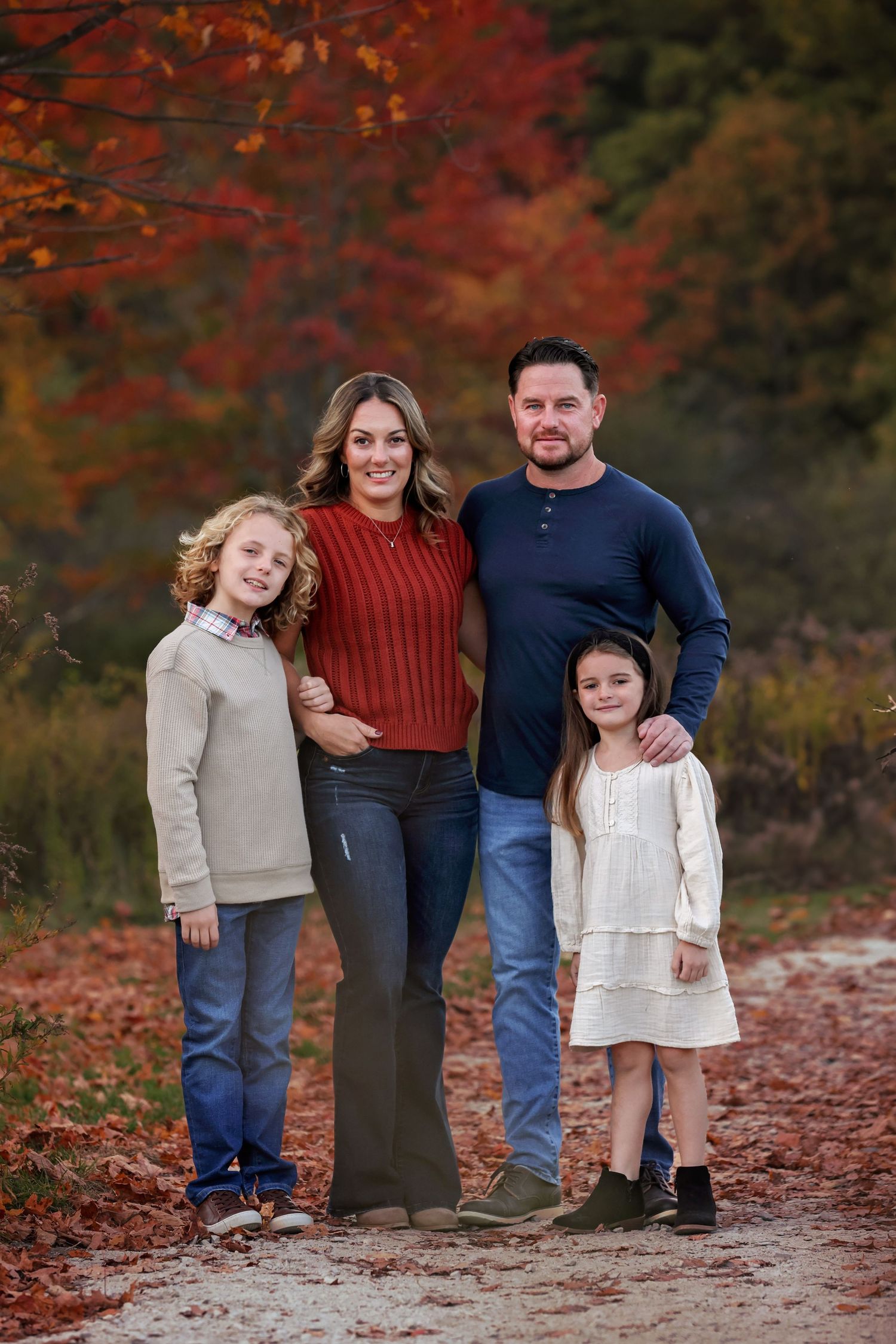 A family poses together on a path surrounded by vibrant red autumn foliage in the background.