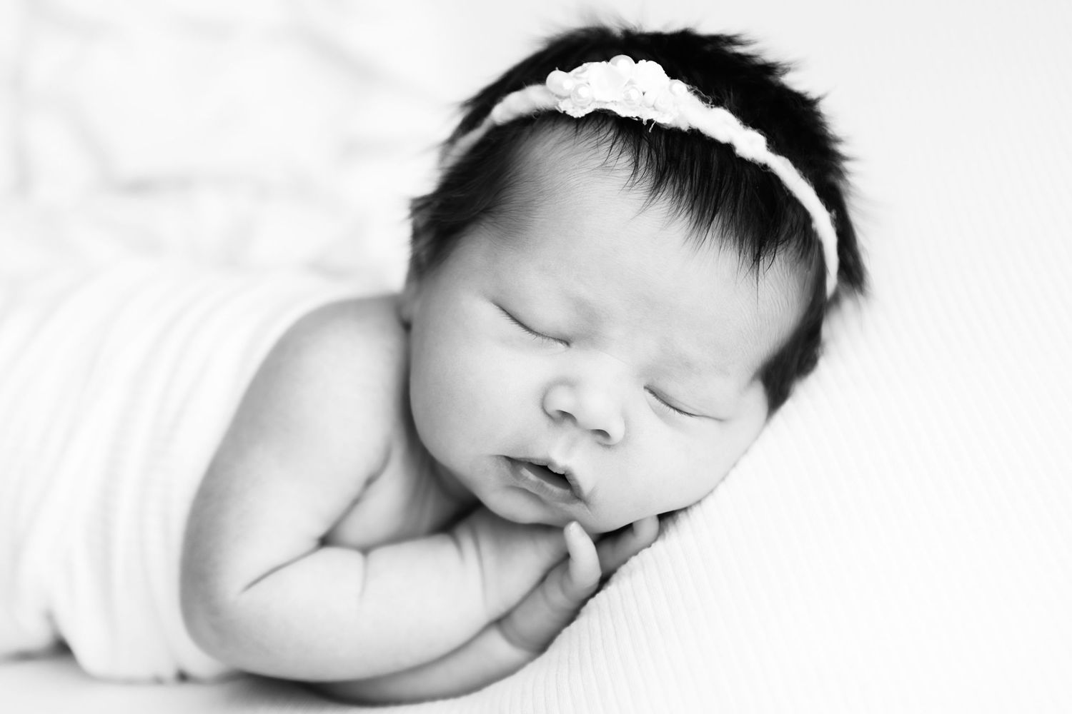 A peaceful sleeping newborn baby wearing a white headband rests on soft white fabric in black and white.