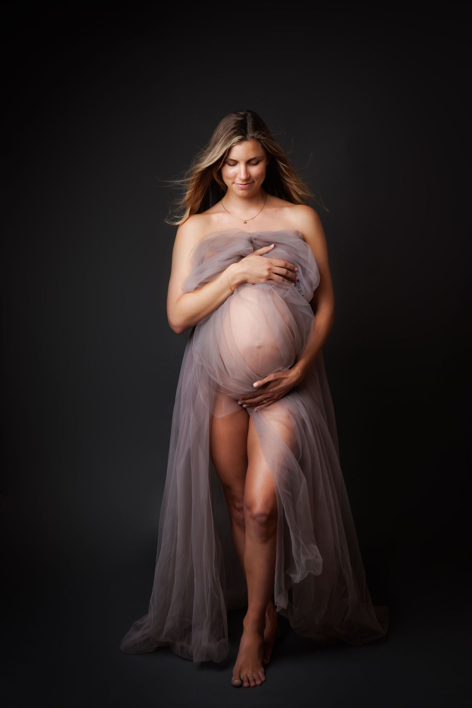 Pregnant woman draped in sheer gray tulle fabric, cradling her baby bump, posing against a dark studio background.