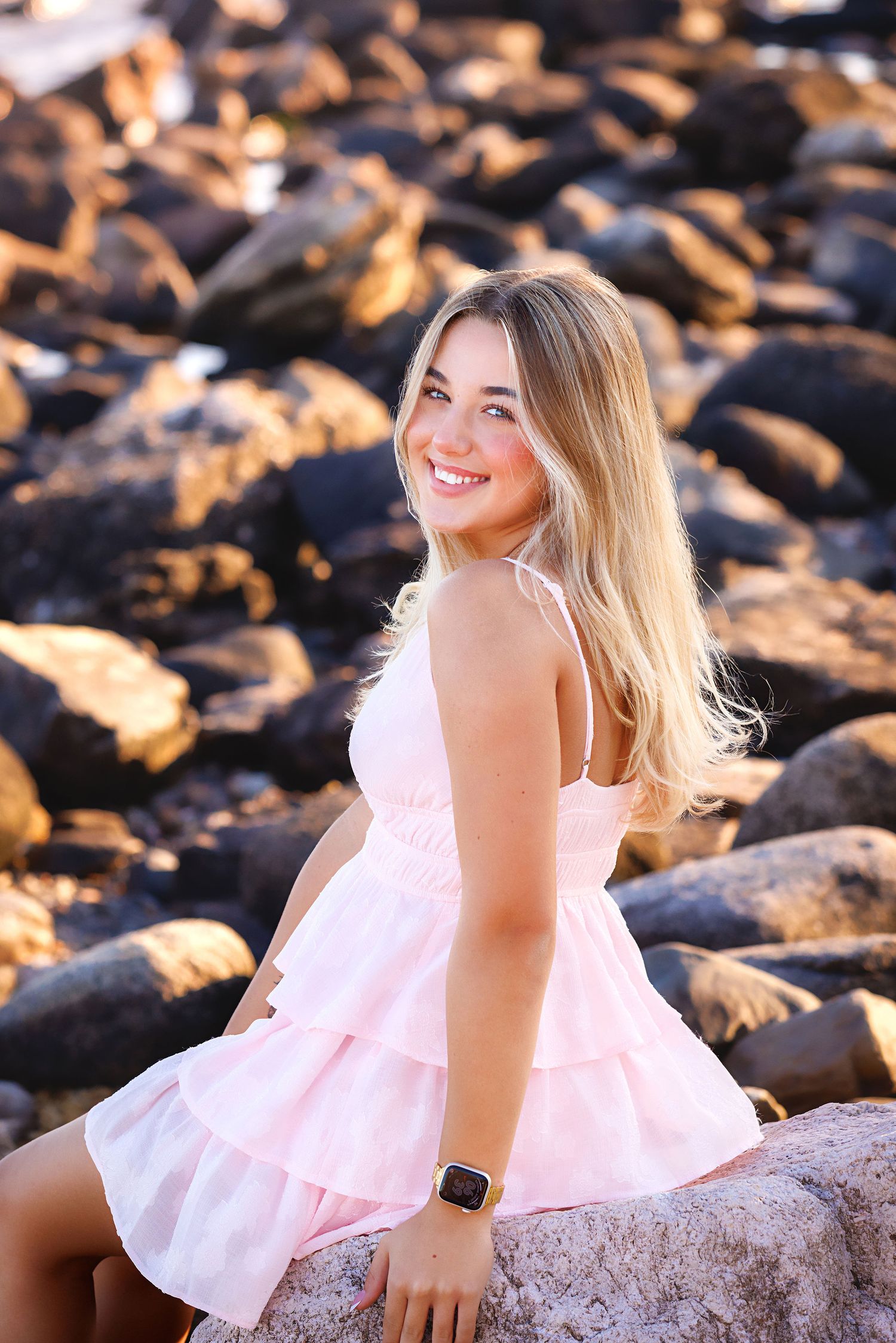 Smiling blonde teen in pink ruffle dress sitting on rocks at beach, wearing a smartwatch, golden hour lighting.