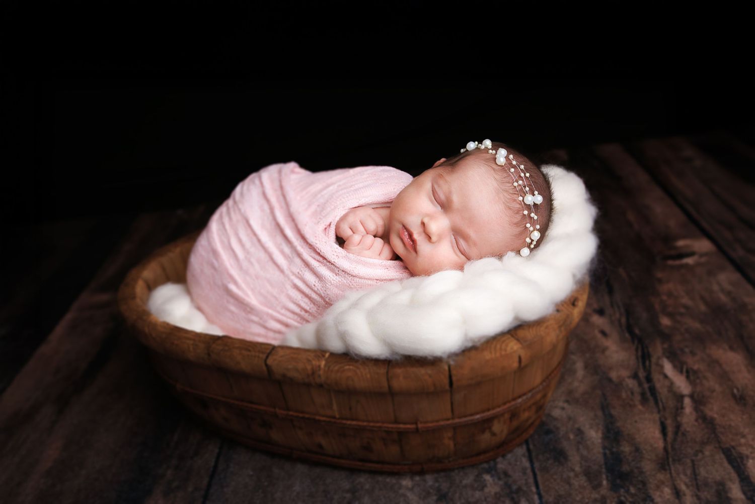 Sleeping newborn baby girl wrapped in pink swaddle with floral headband resting in wooden bowl on rustic surface.