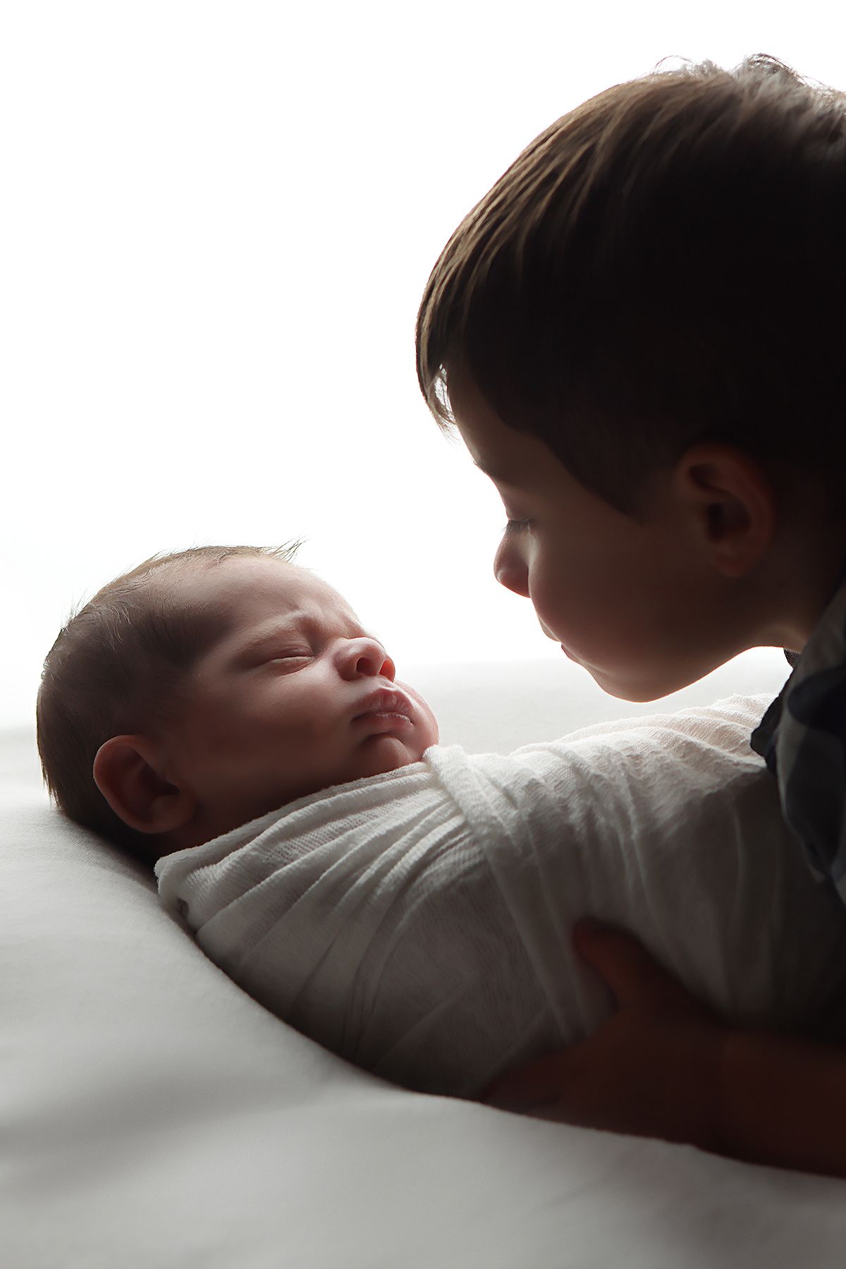 A tender moment between siblings as an older child looks lovingly at a swaddled newborn baby lying on white bedding.