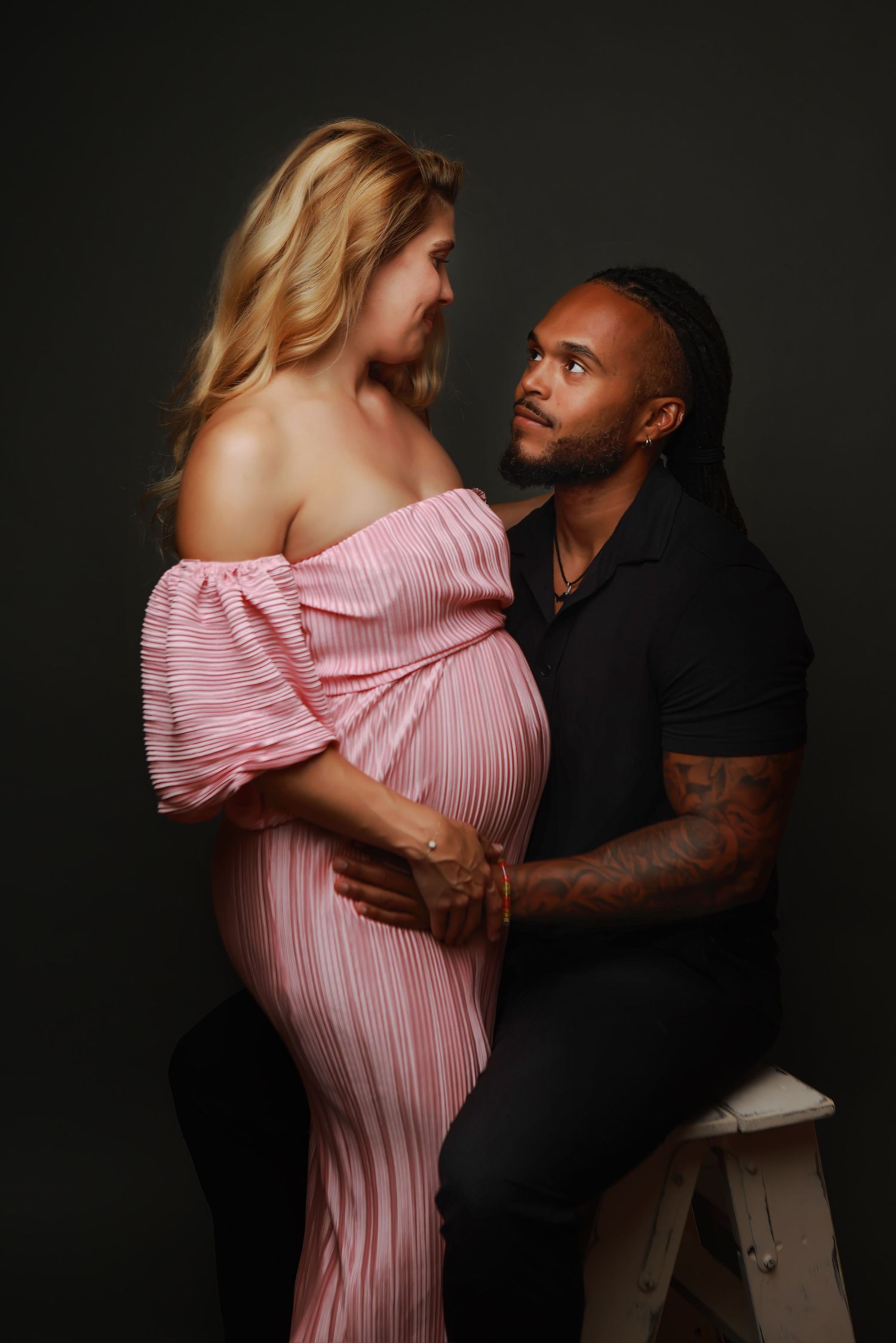 A couple in a romantic embrace poses for a series of intimate maternity photos against a dark background.