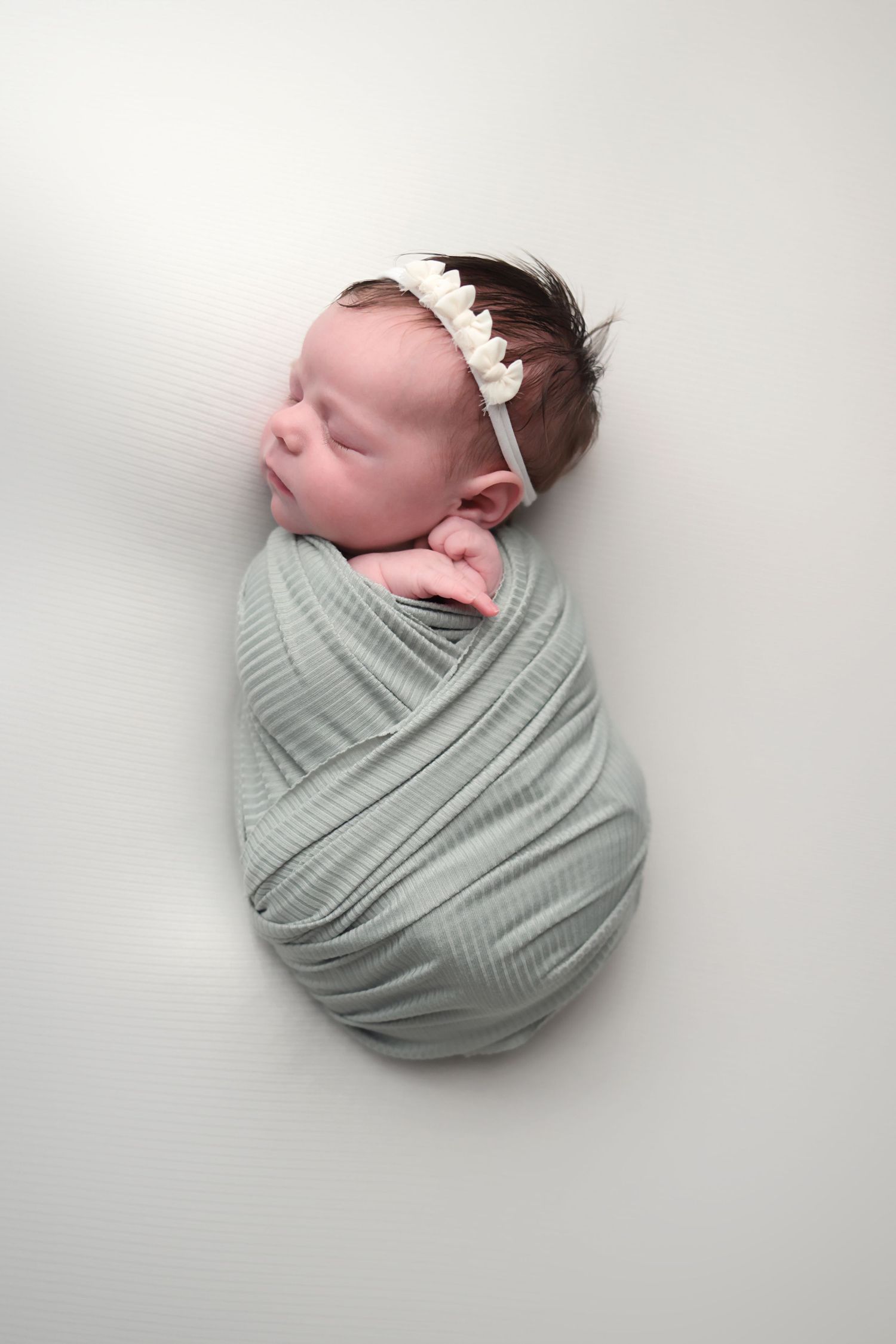 A sleeping newborn baby swaddled in a soft sage green wrap against a white background.